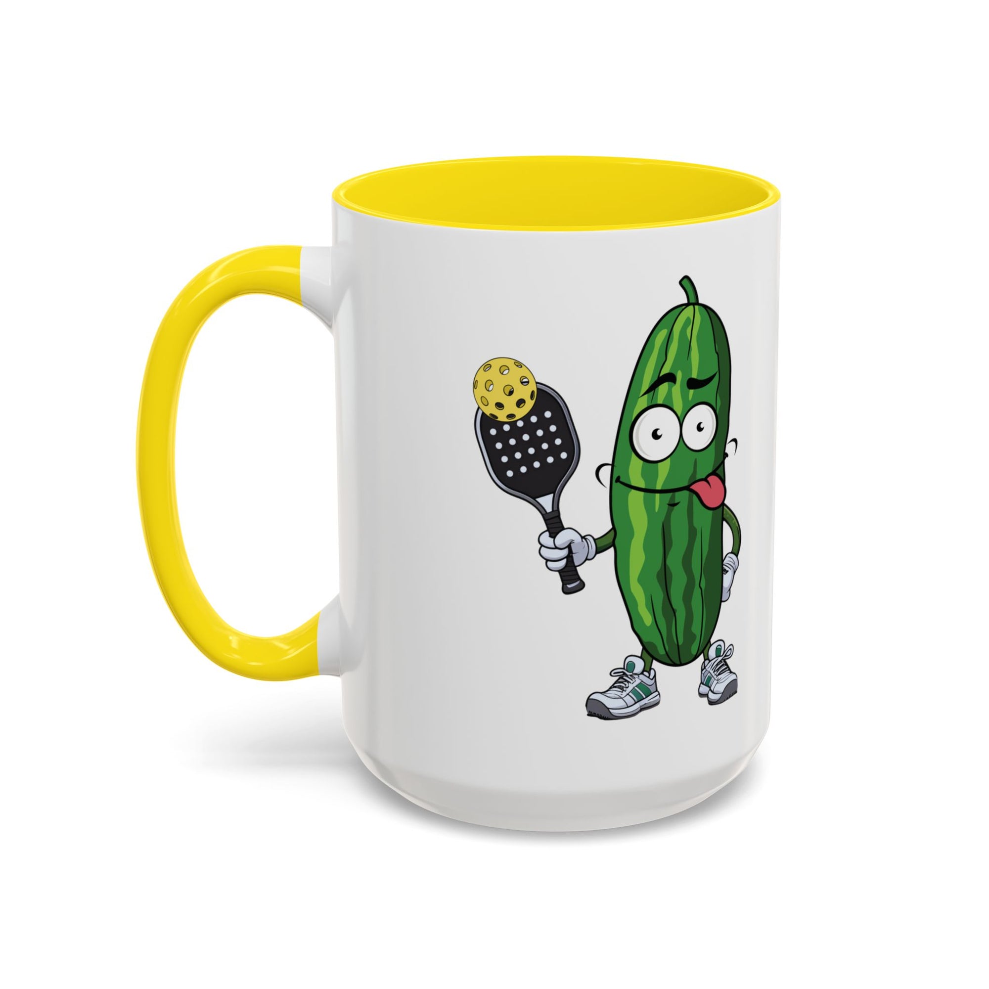 A ceramic coffee mug with yellow accents on the handle and interior, with a cartoon pickle with googly eyes and his tongue sticking out, holding a pickleball paddle with a yellow pickleball, against a white background