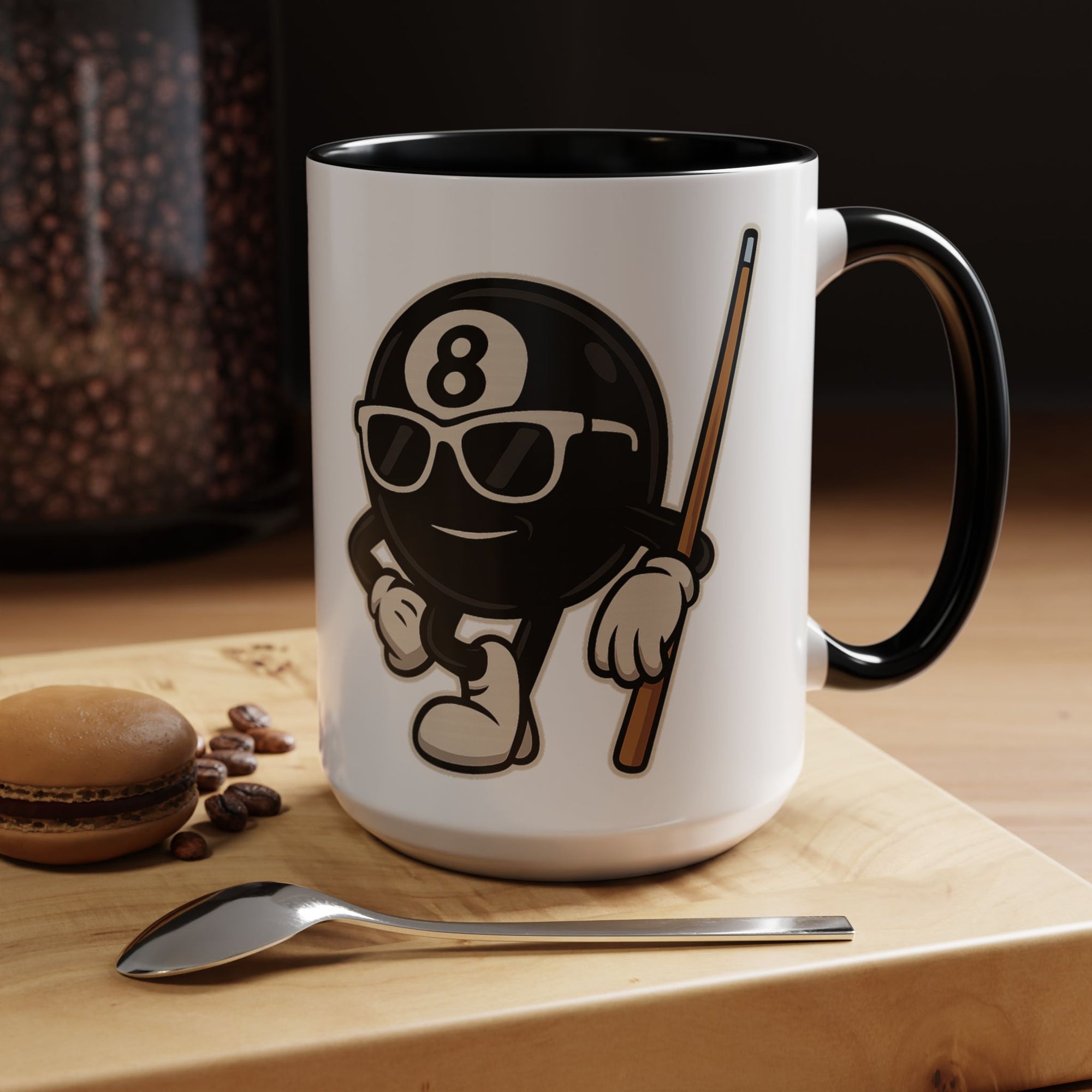 A white ceramic coffee mug with black accents on the handle and interior, with a graphic of a cartoon billiards 8 ball with arms and legs and wearing sunglasses and leaning on a cue stick, in a coffee shop setting