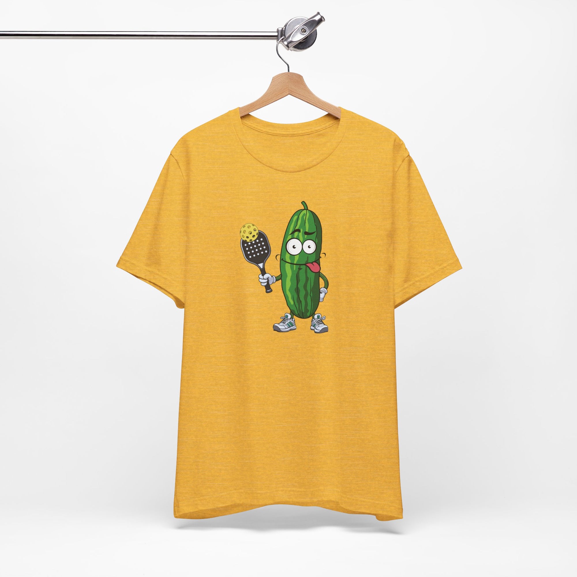 Yellow t-shirt with a cartoon pickle with googly eyes and his tongue sticking out, holding a pickleball paddle with a yellow pickleball, on a hanger against a white background