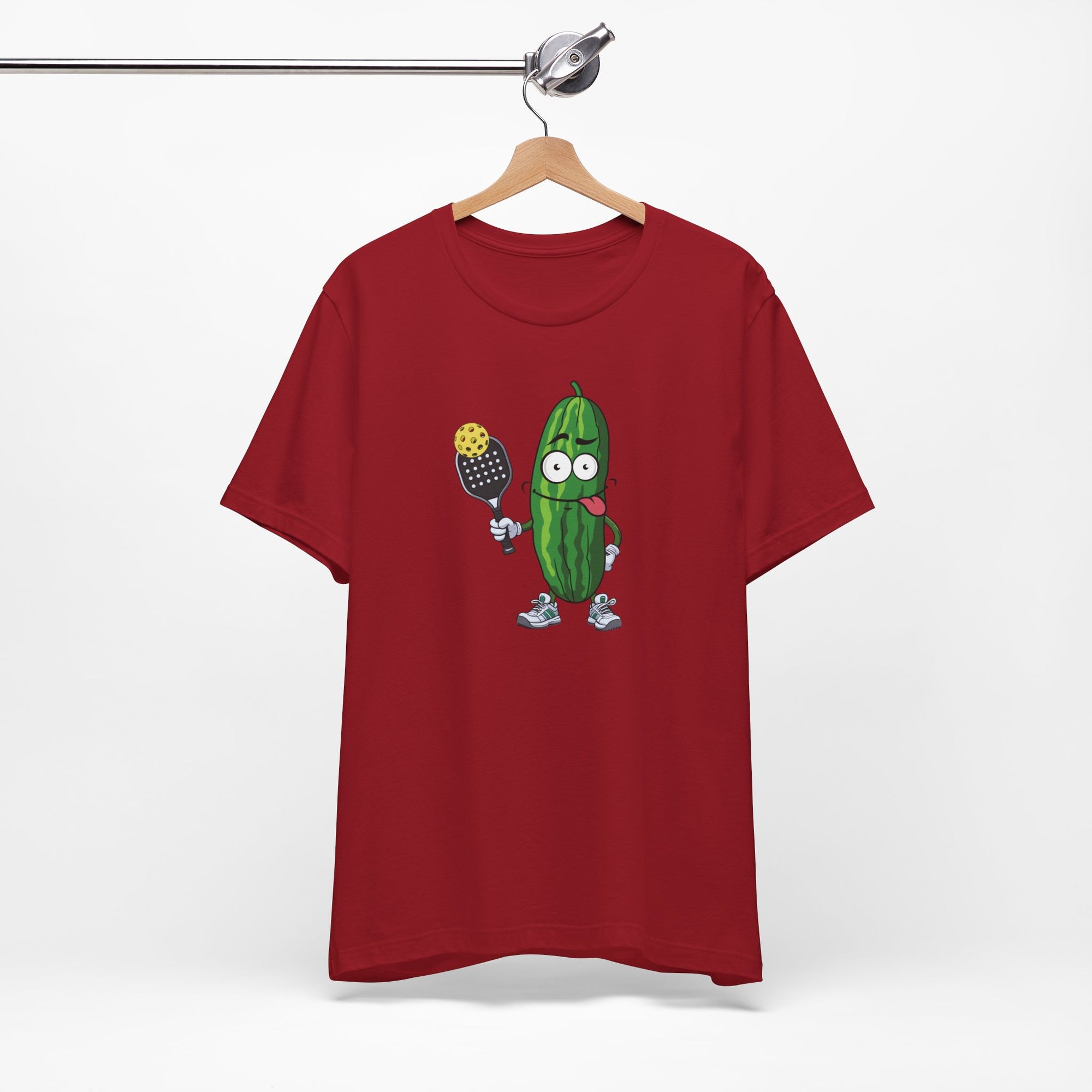 Red t-shirt with a cartoon pickle with googly eyes and his tongue sticking out, holding a pickleball paddle with a yellow pickleball, on a hanger against a white background
