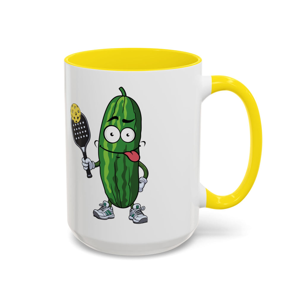 A ceramic coffee mug with yellow accents on the handle and interior, with a cartoon pickle with googly eyes and his tongue sticking out, holding a pickleball paddle with a yellow pickleball, against a white background
