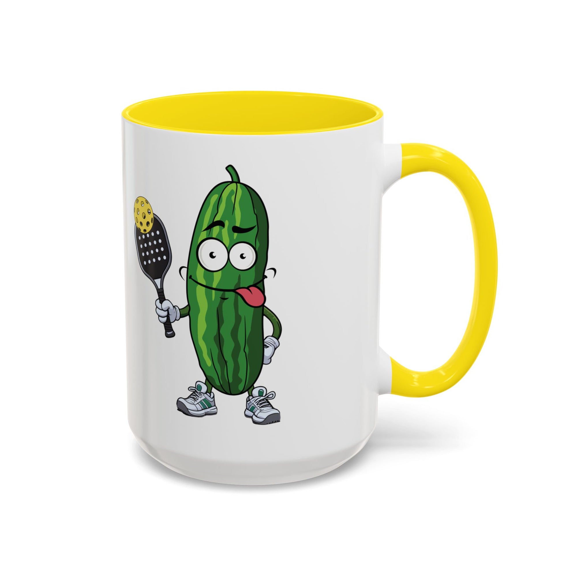 A ceramic coffee mug with yellow accents on the handle and interior, with a cartoon pickle with googly eyes and his tongue sticking out, holding a pickleball paddle with a yellow pickleball, against a white background