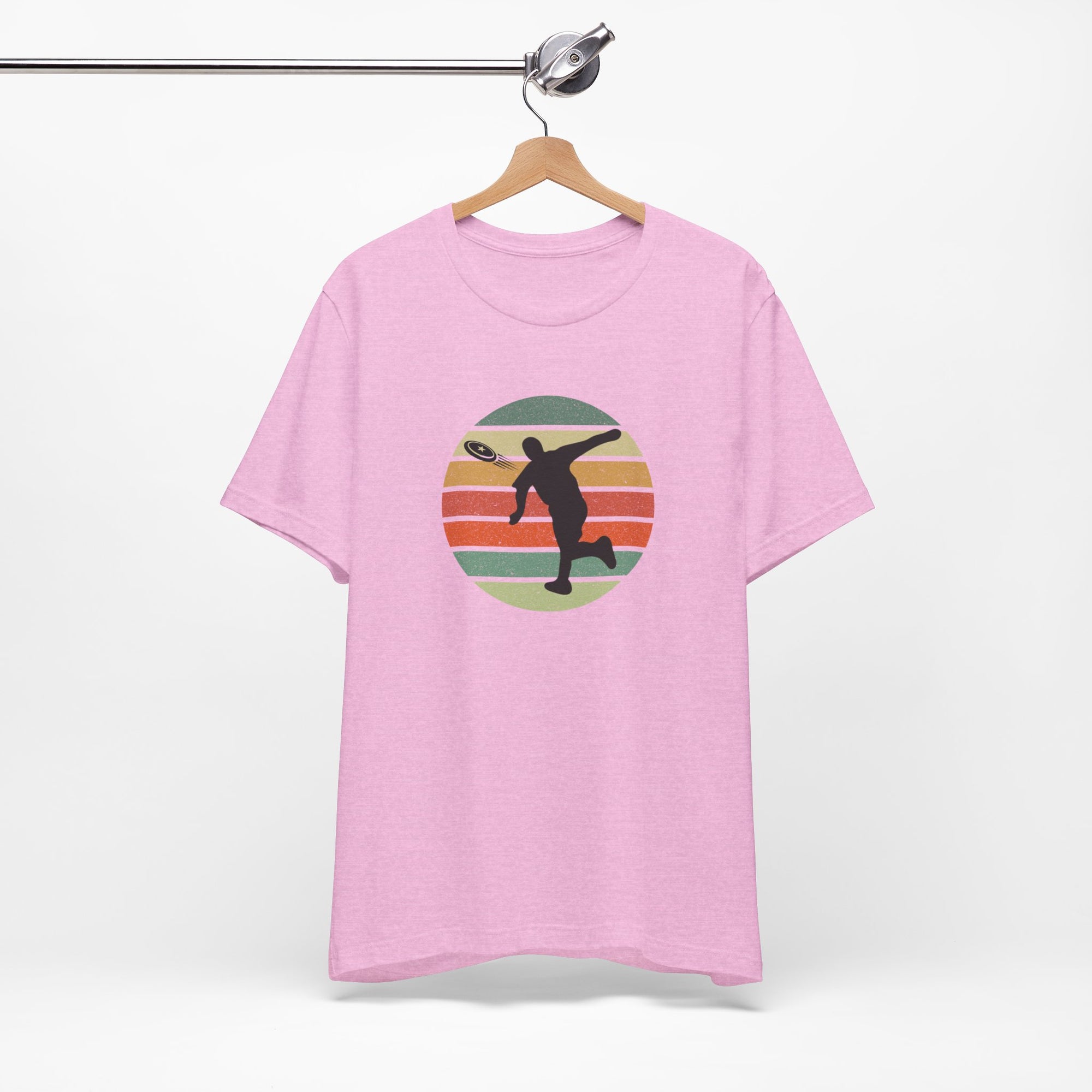 Pink t-shirt with a graphic of a silhouette disc golf player throwing a disc above a colorful retro graphic, hanging on a wooden hanger.
