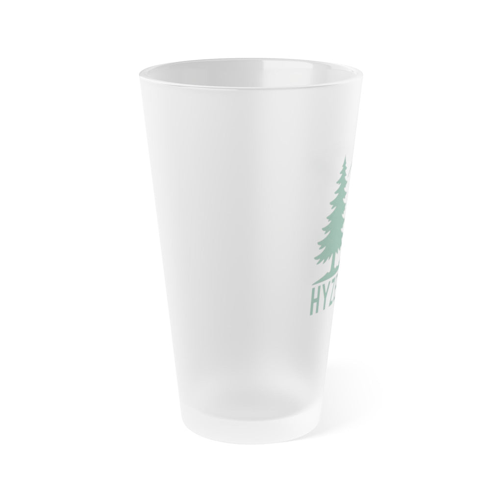 Side view of a frosted pint glass with a design that says Hyzer Life with a flying disc golf disc, basket, and chains along with trees in the background.