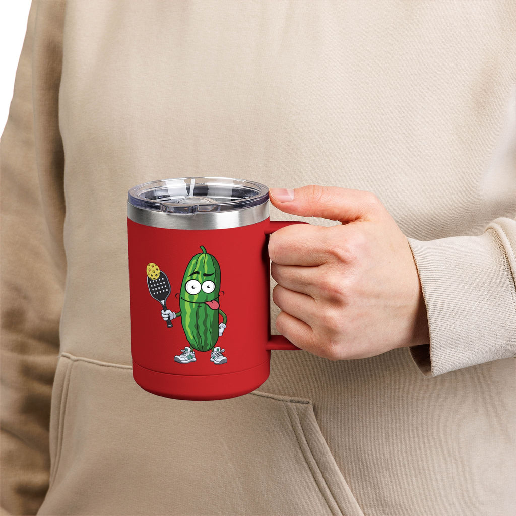A person holding a red travel coffee mug tumbler with handle and lid with a cartoon pickle with googly eyes and his tongue sticking out, holding a pickleball paddle with a yellow pickleball