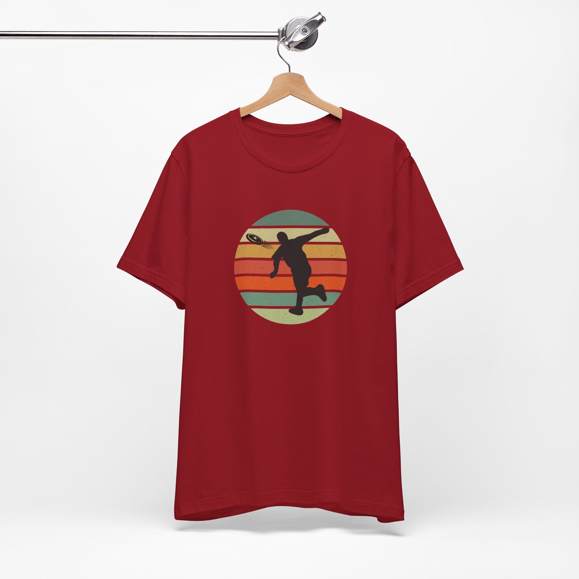Red t-shirt with a graphic of a silhouette disc golf player throwing a disc above a colorful retro graphic, hanging on a wooden hanger.