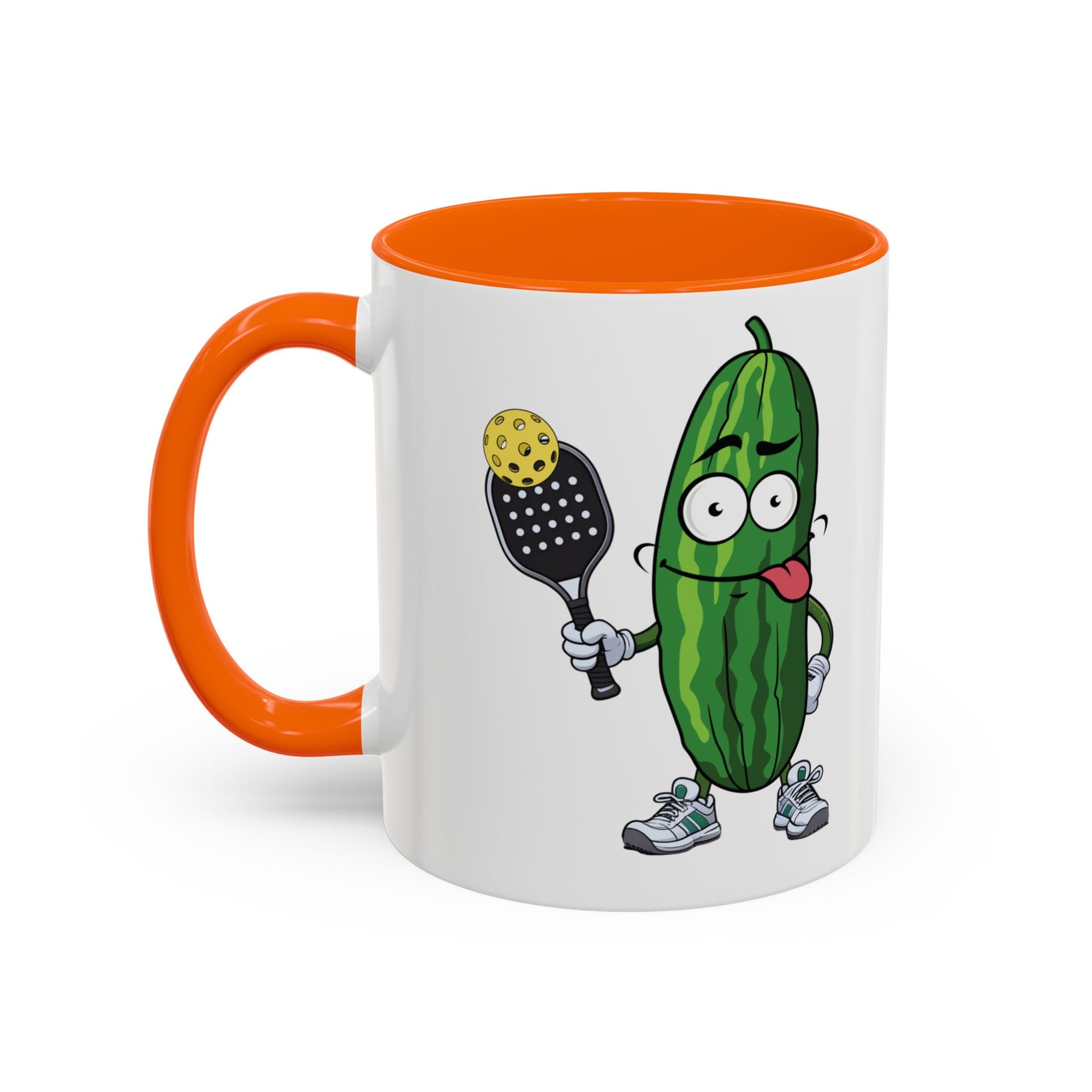 A ceramic coffee mug with orange accents on the handle and interior, with a cartoon pickle with googly eyes and his tongue sticking out, holding a pickleball paddle with a yellow pickleball, against a white background