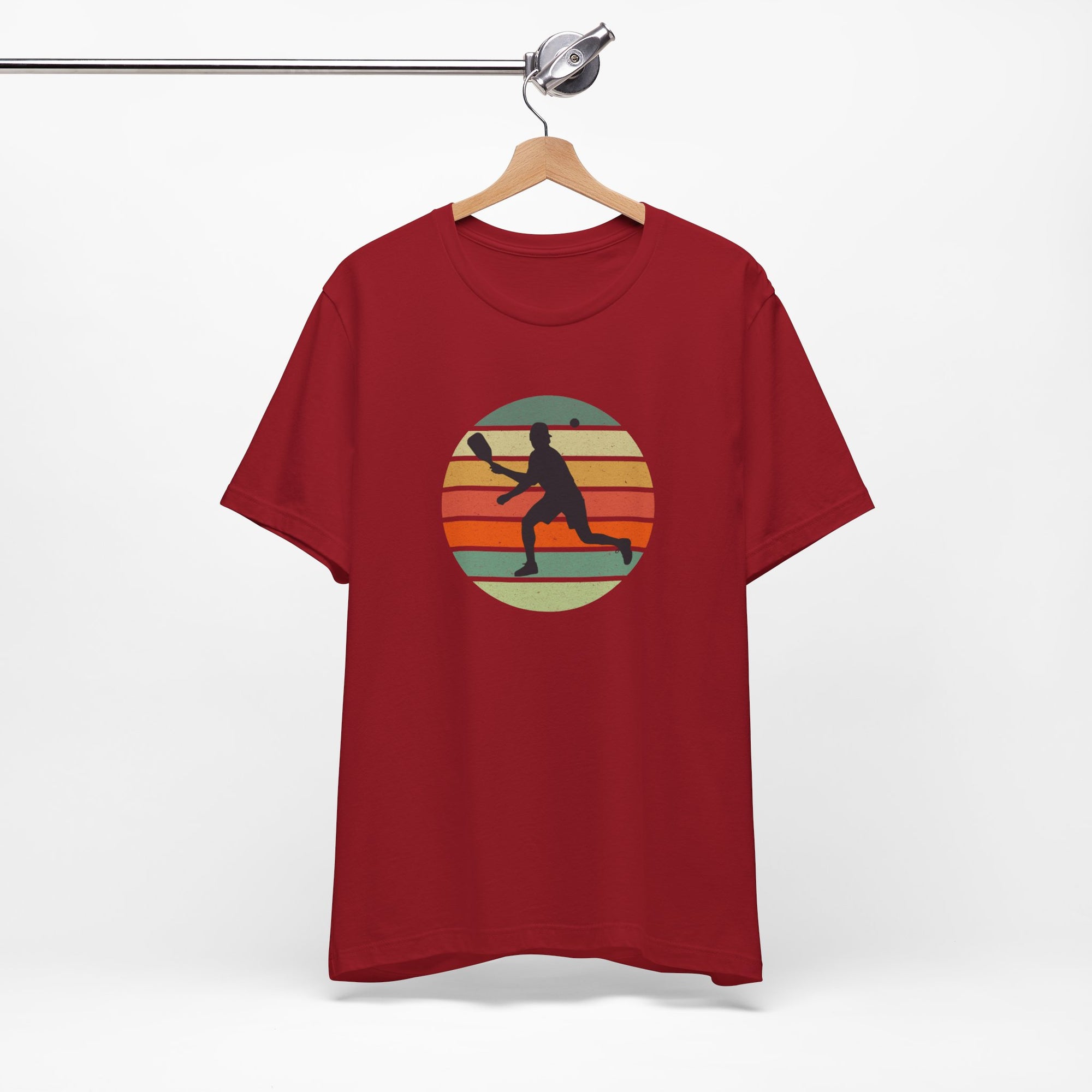 Red t-shirt with a graphic design of a person playing pickleball against a retro colorful background, hanging on a wooden hanger.