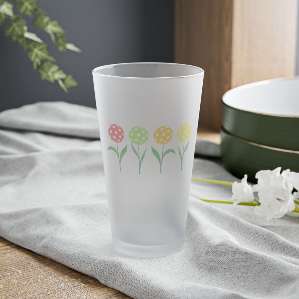 A frosted pint glass with four colorful flowers made out of pickleballs, on a tabletop.