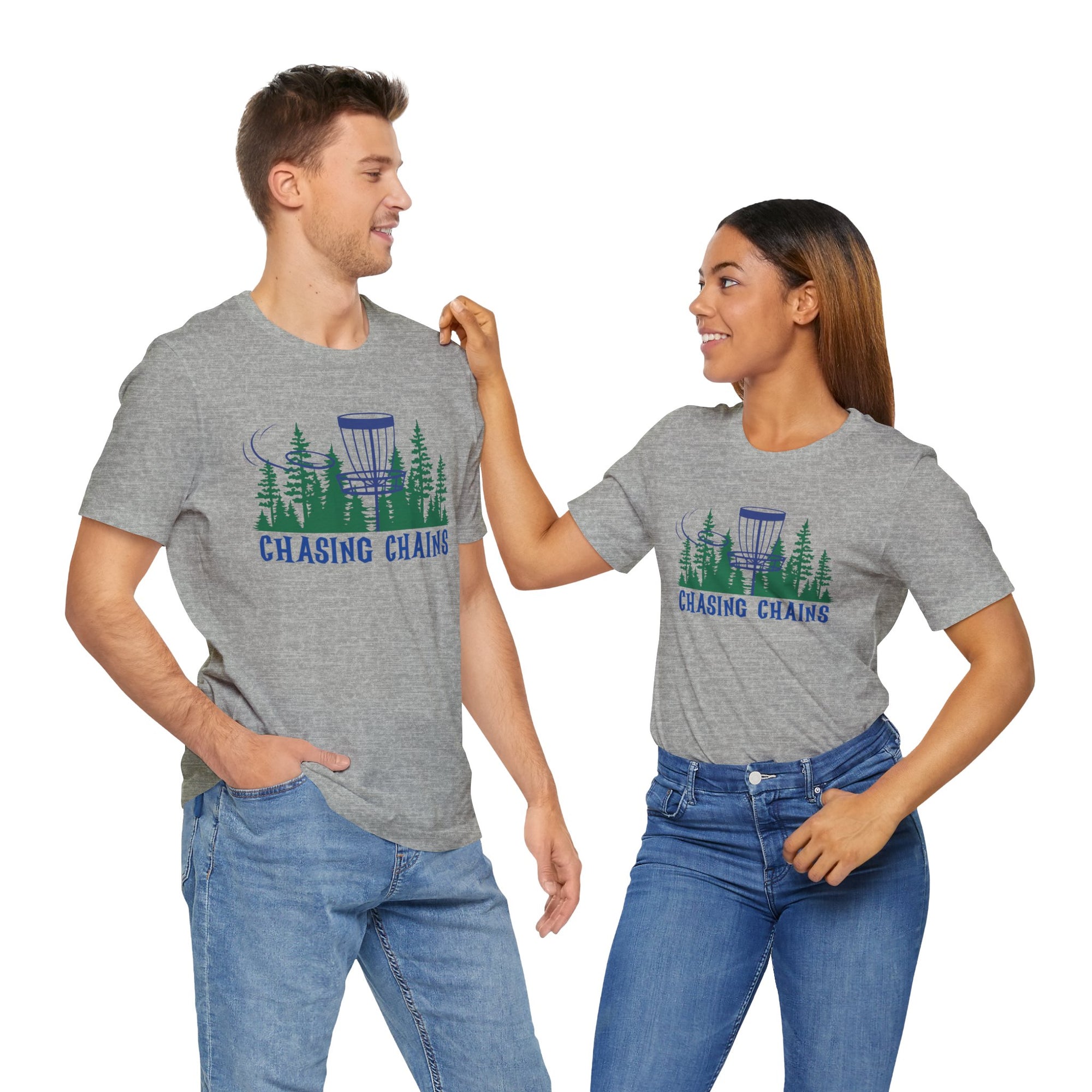 A man and a woman both wearing a gray t-shirt with 'Chasing Chains' graphic featuring a flying disc golf disc and basket and chains in front of a scenic evergreen tree landscape, against a white background