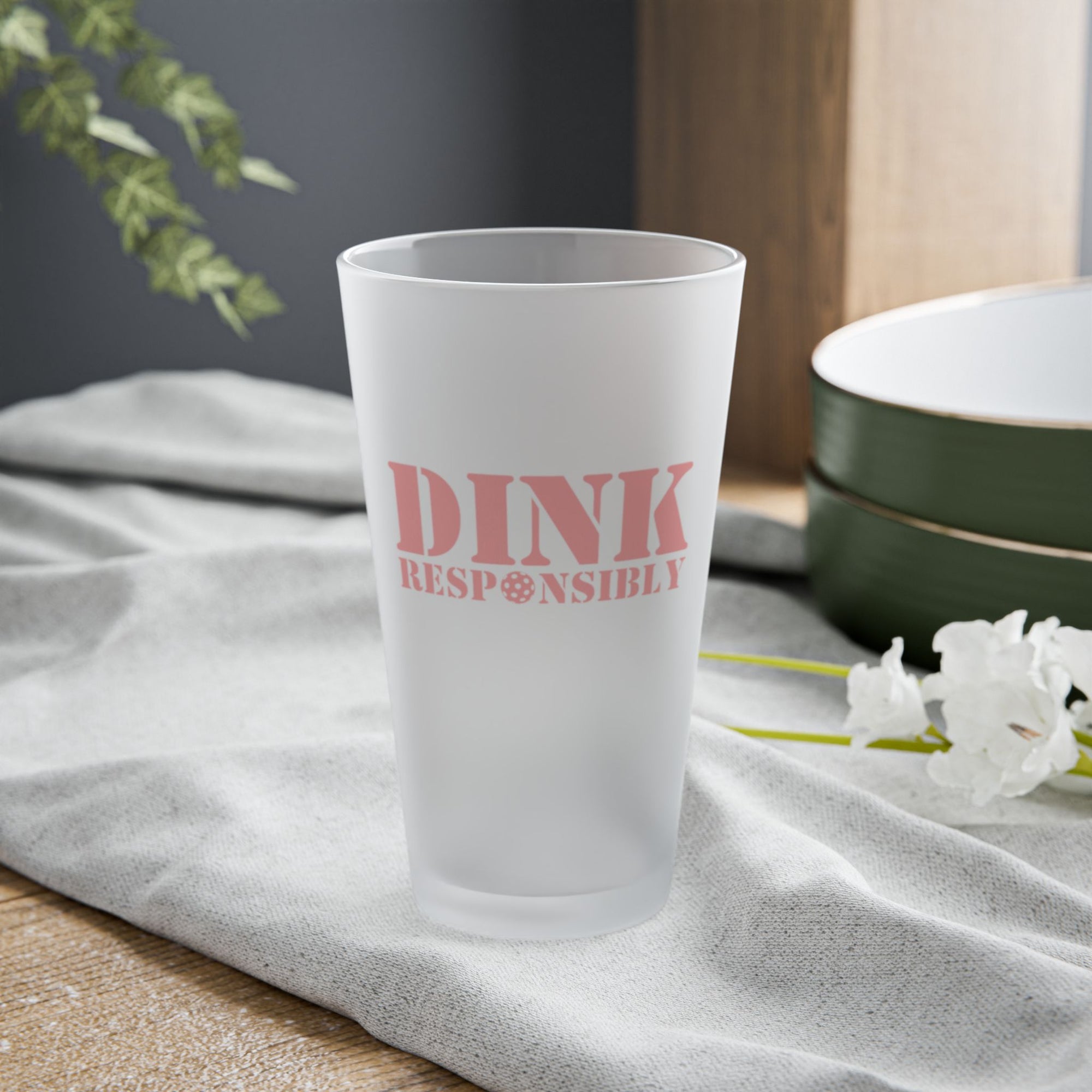 A frosted pint glass with "Dink Responsibly" with a pickleball replacing the "O" in "Responsibly," on a tabletop.