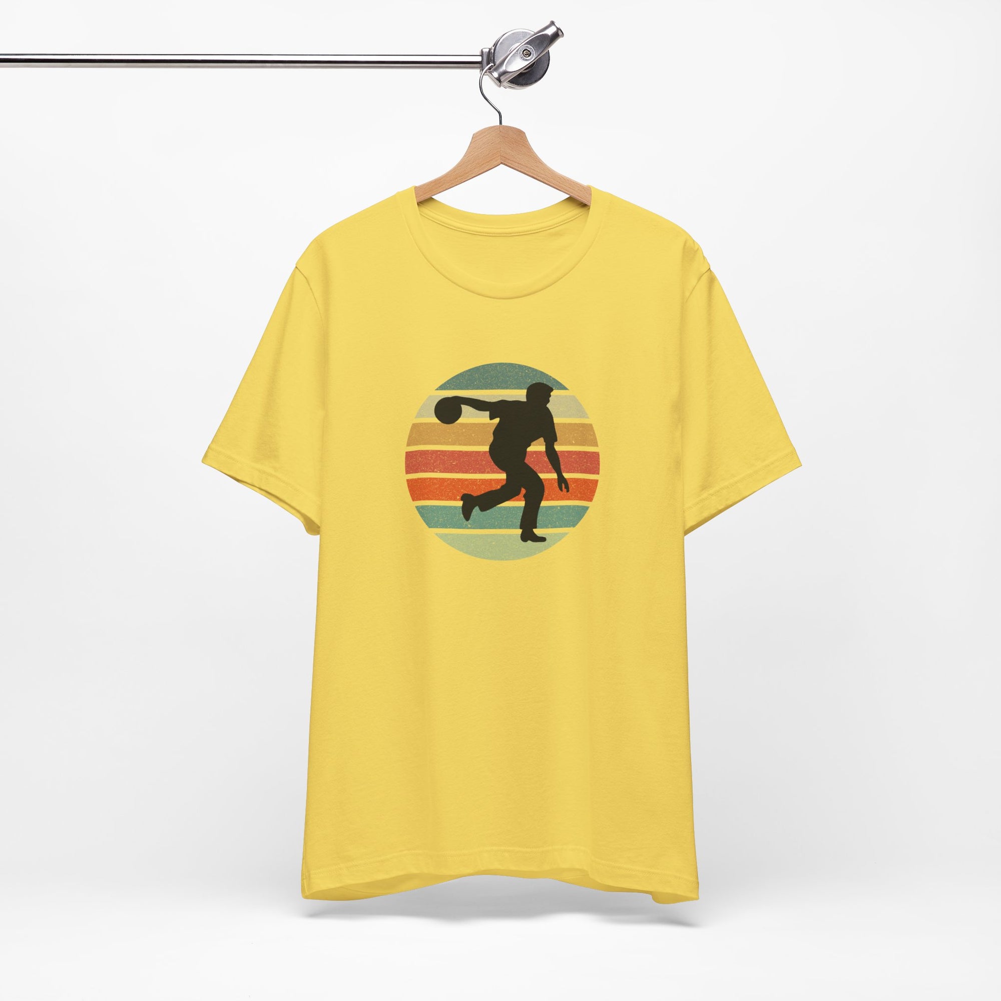 Yellow t-shirt with a graphic of a silhouette of a bowler over a colorful retro image, hanging on a wooden hanger against a white background.