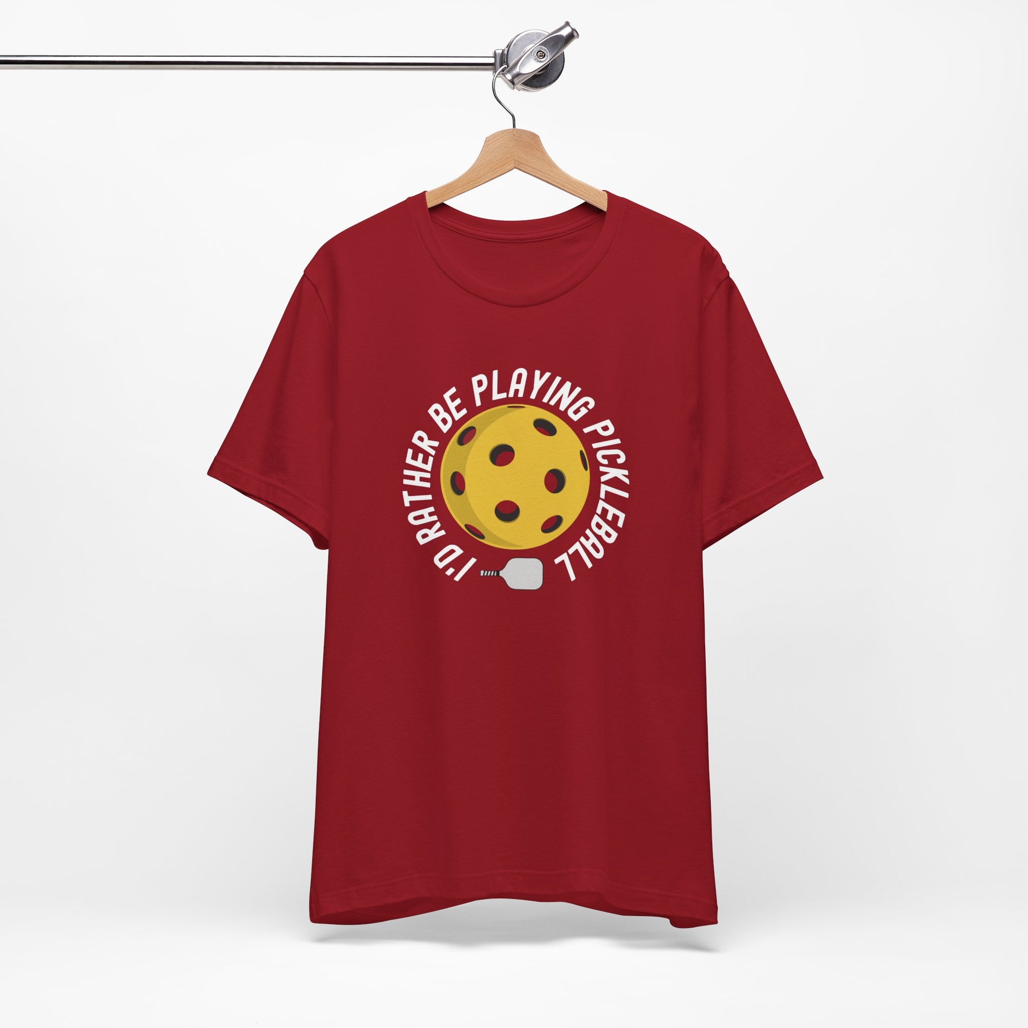 Red t-shirt with a yellow pickleball and white pickleball paddle and the text "I'd Rather Be Playing Pickleball" on a hanger against a white background