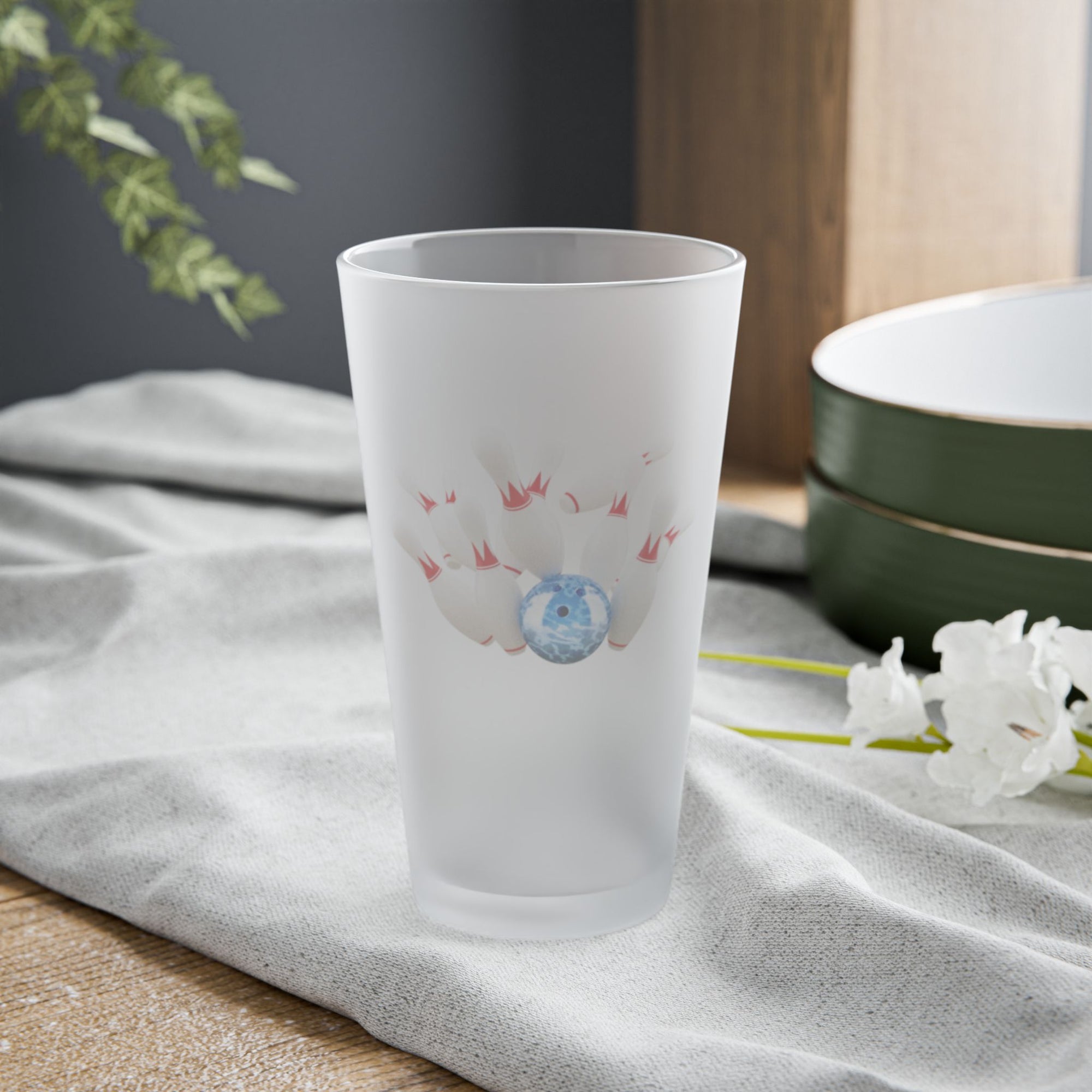A frosted pint glass with a graphic of a blue bowling ball knocking over bowling pins, on a tabletop