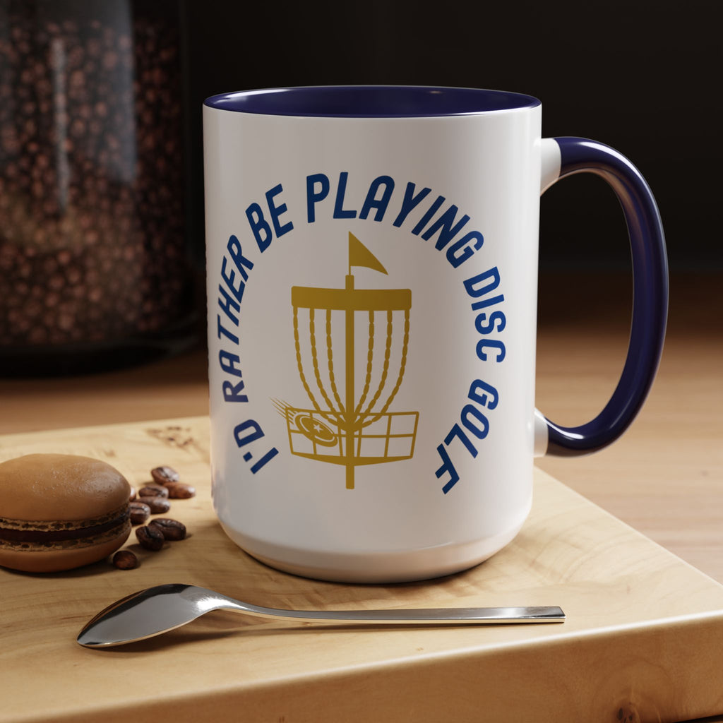 Ceramic coffee mug with blue accents on the handle and inside, with a graphic of disc golf chains, basket, and disc and text that says "I'd Rather Be Playing Disc Golf" in a coffee shop setting.