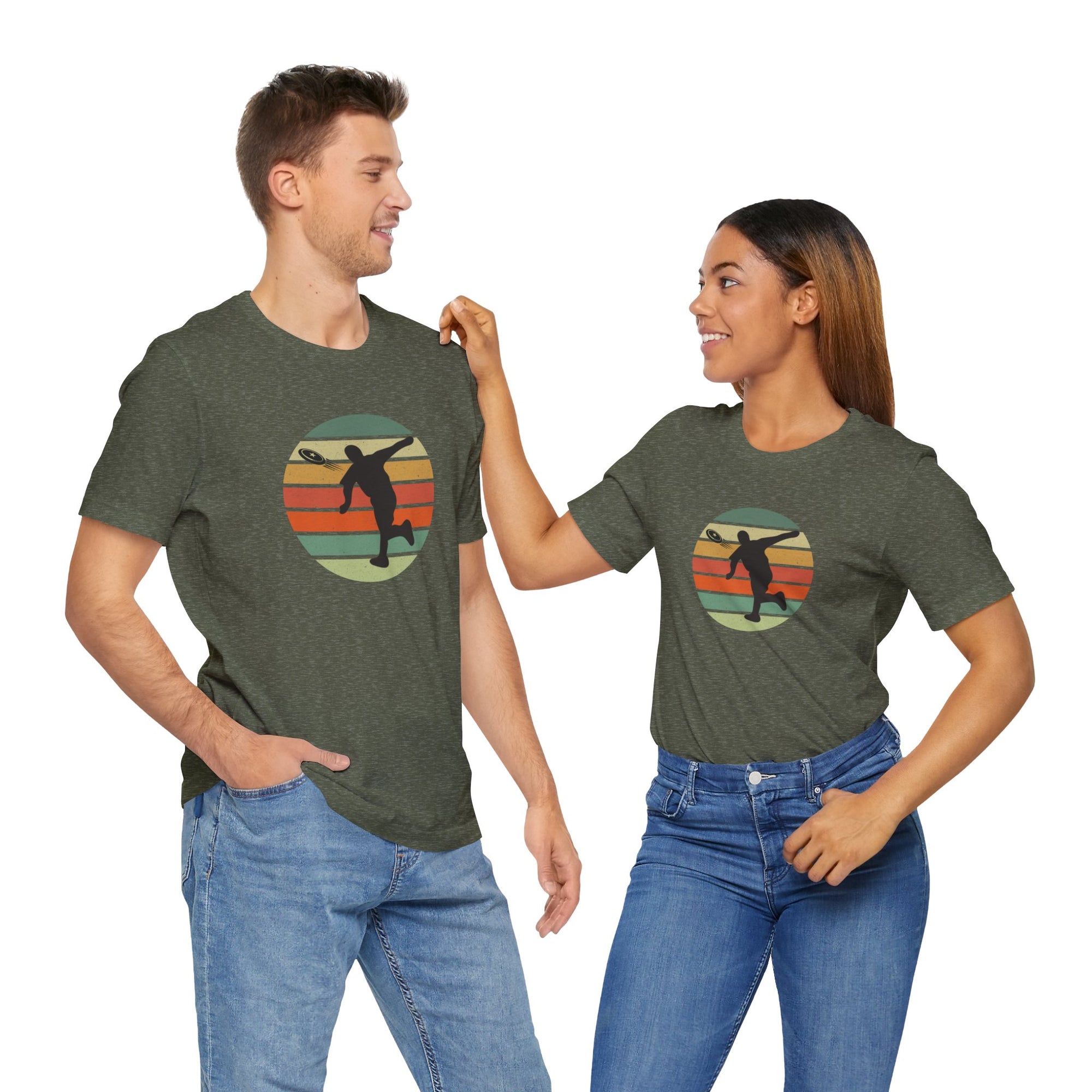 A man and a woman both wearing amilitary green t-shirt with a graphic of a silhouette disc golf player throwing a disc above a colorful retro graphic, hanging on a wooden hanger.
