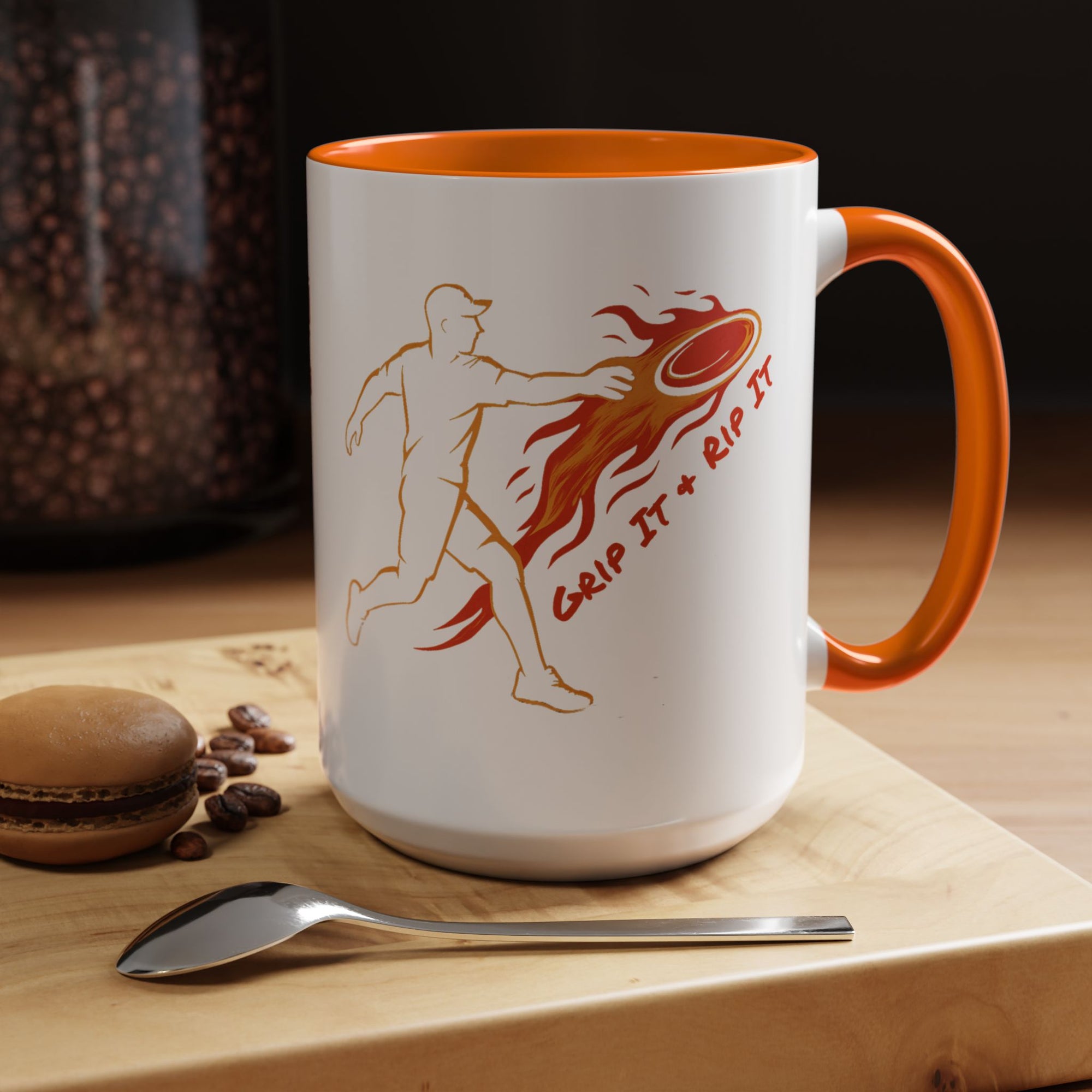 A ceramic coffee mug with orange accents on the handle and interior, featuring a graphic design of a person throwing a disc golf disc with flames coming off the disc and text 'Grip It or Rip It' in a coffee shop setting.