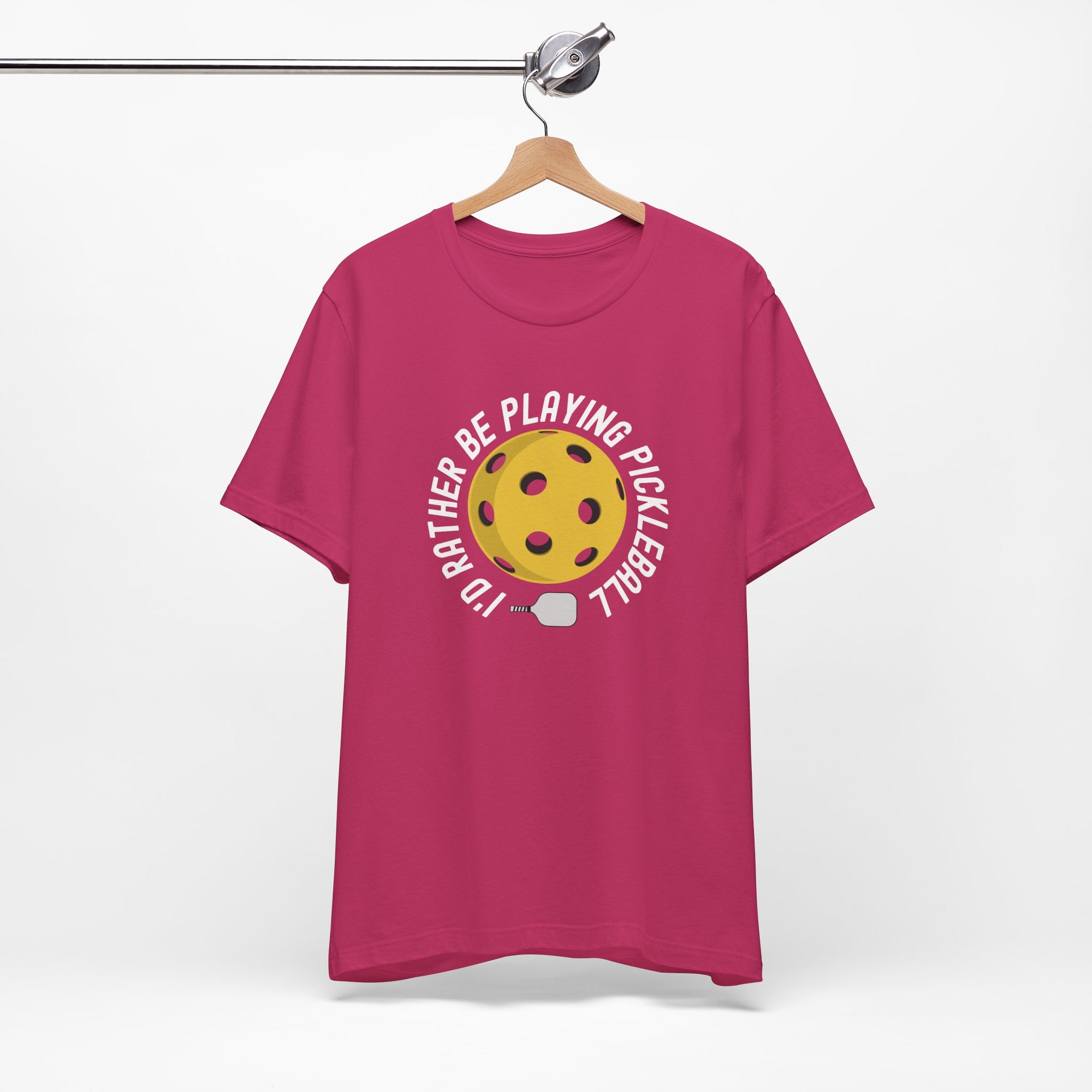 Pink t-shirt with a yellow pickleball and white pickleball paddle and the text "I'd Rather Be Playing Pickleball" on a hanger against a white background