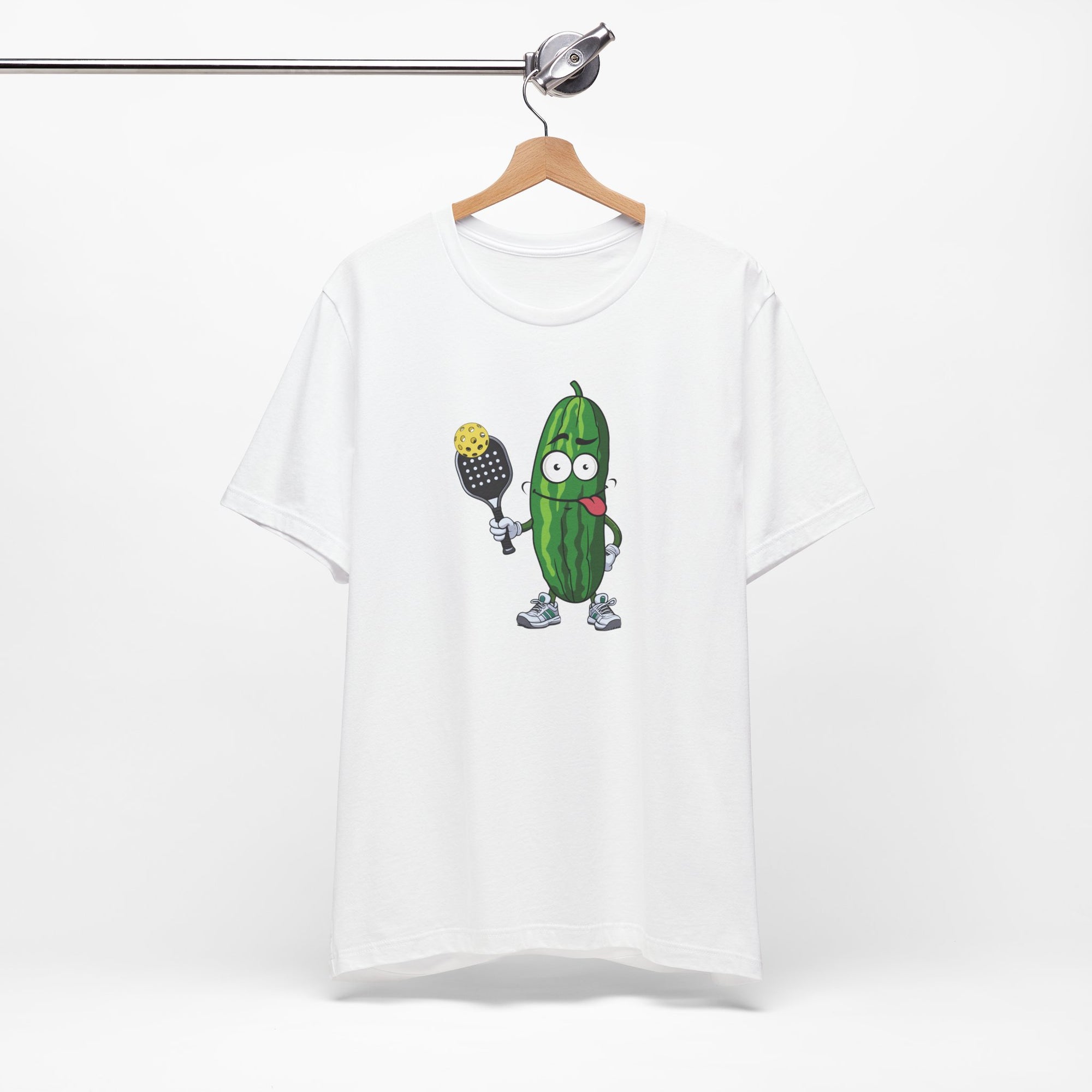 White t-shirt with a cartoon pickle with googly eyes and his tongue sticking out, holding a pickleball paddle with a yellow pickleball, on a hanger against a white background