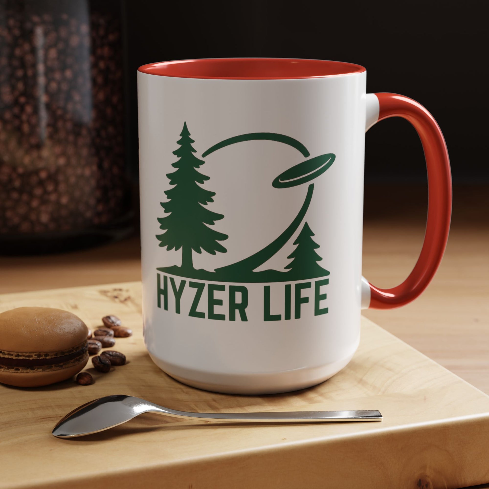 Ceramic coffee mug with red accents on the handle and interior, with green graphic of trees and a disc golf flying with the words 'HYZER LIFE' below the image, in a coffee shop setting.