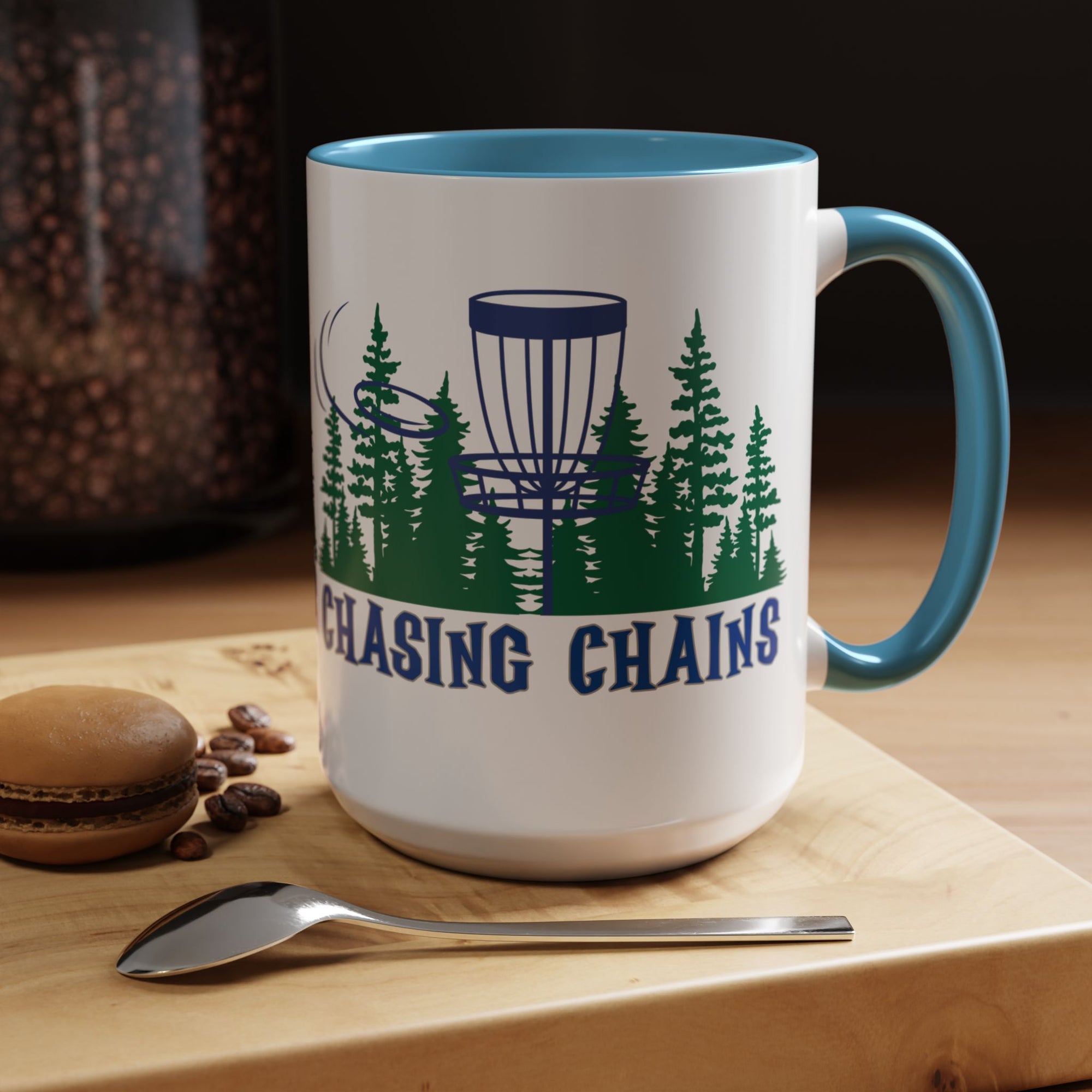 A ceramic coffee mug with blue accents on the handle and interior, with 'Chasing Chains' graphic featuring a flying disc golf disc and basket and chains in front of a scenic evergreen tree landscape, in a coffee shop setting