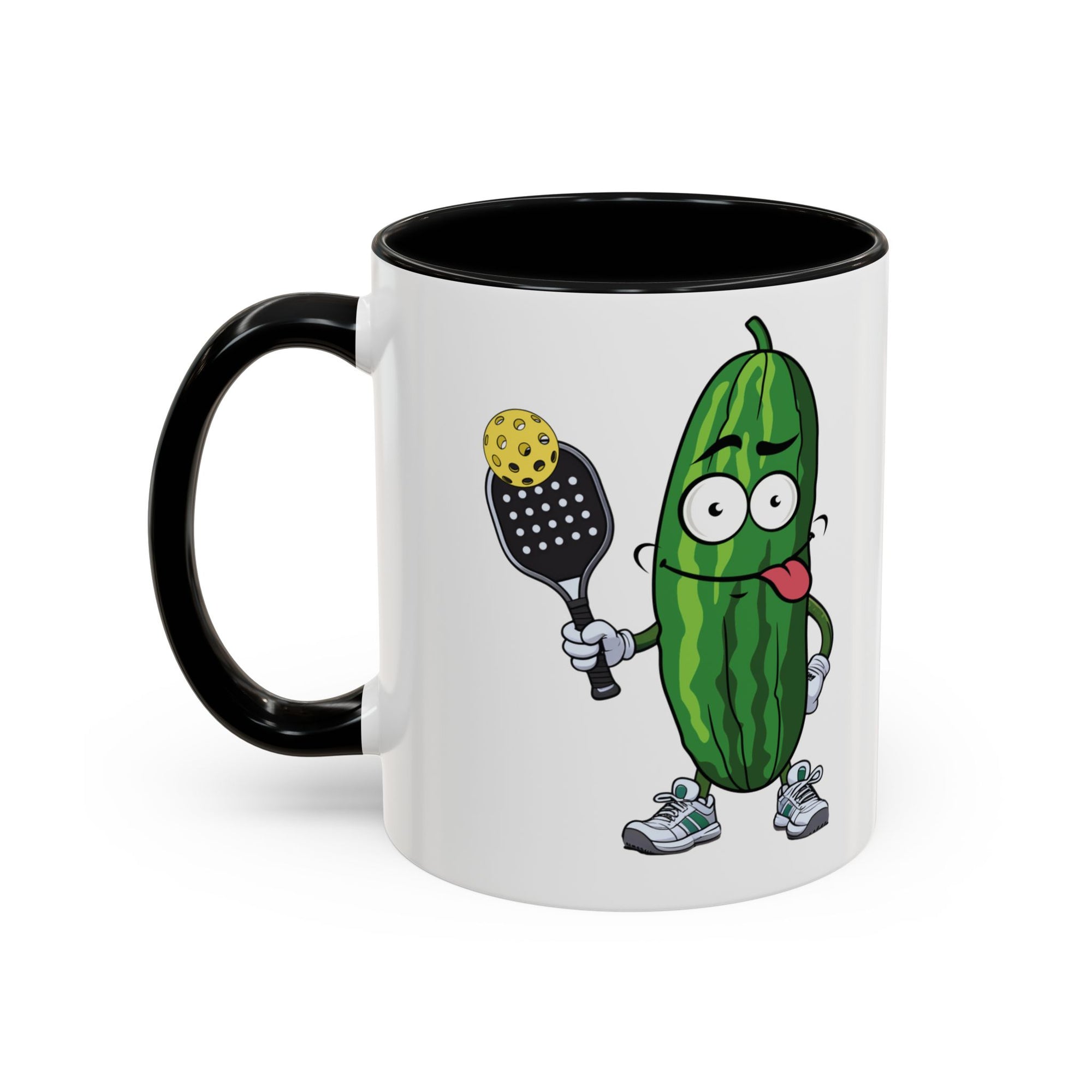 A ceramic coffee mug with black accents on the handle and interior, with a cartoon pickle with googly eyes and his tongue sticking out, holding a pickleball paddle with a yellow pickleball, against a white background