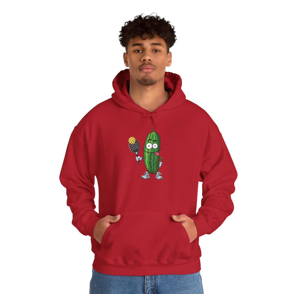 A man wearing a red hoodie with a cartoon pickle with googly eyes and his tongue sticking out, holding a pickleball paddle with a yellow pickleball, against a white background