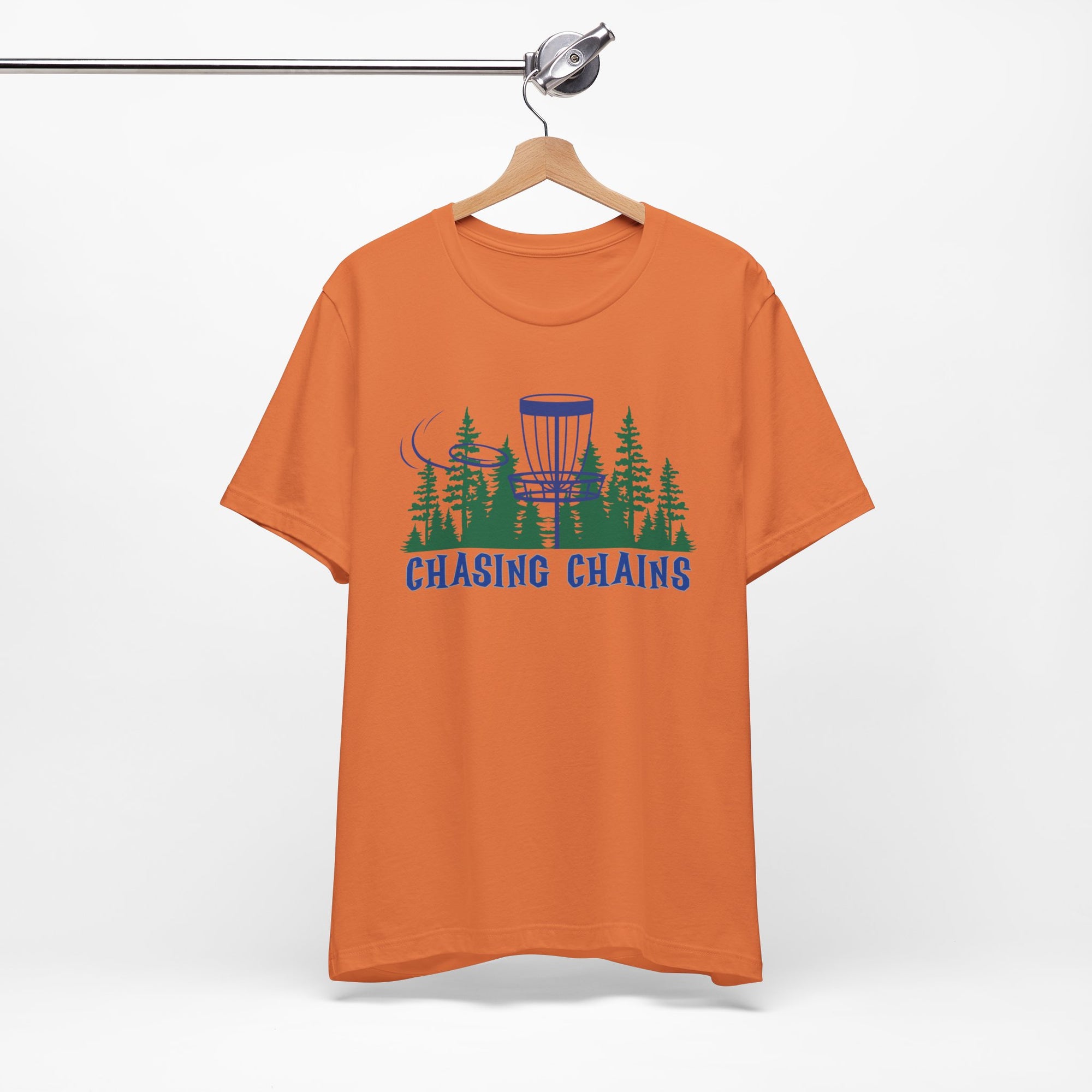Orange t-shirt with 'Chasing Chains' graphic featuring a flying disc golf disc and basket and chains in front of a scenic evergreen tree landscape, on a hanger against a white background