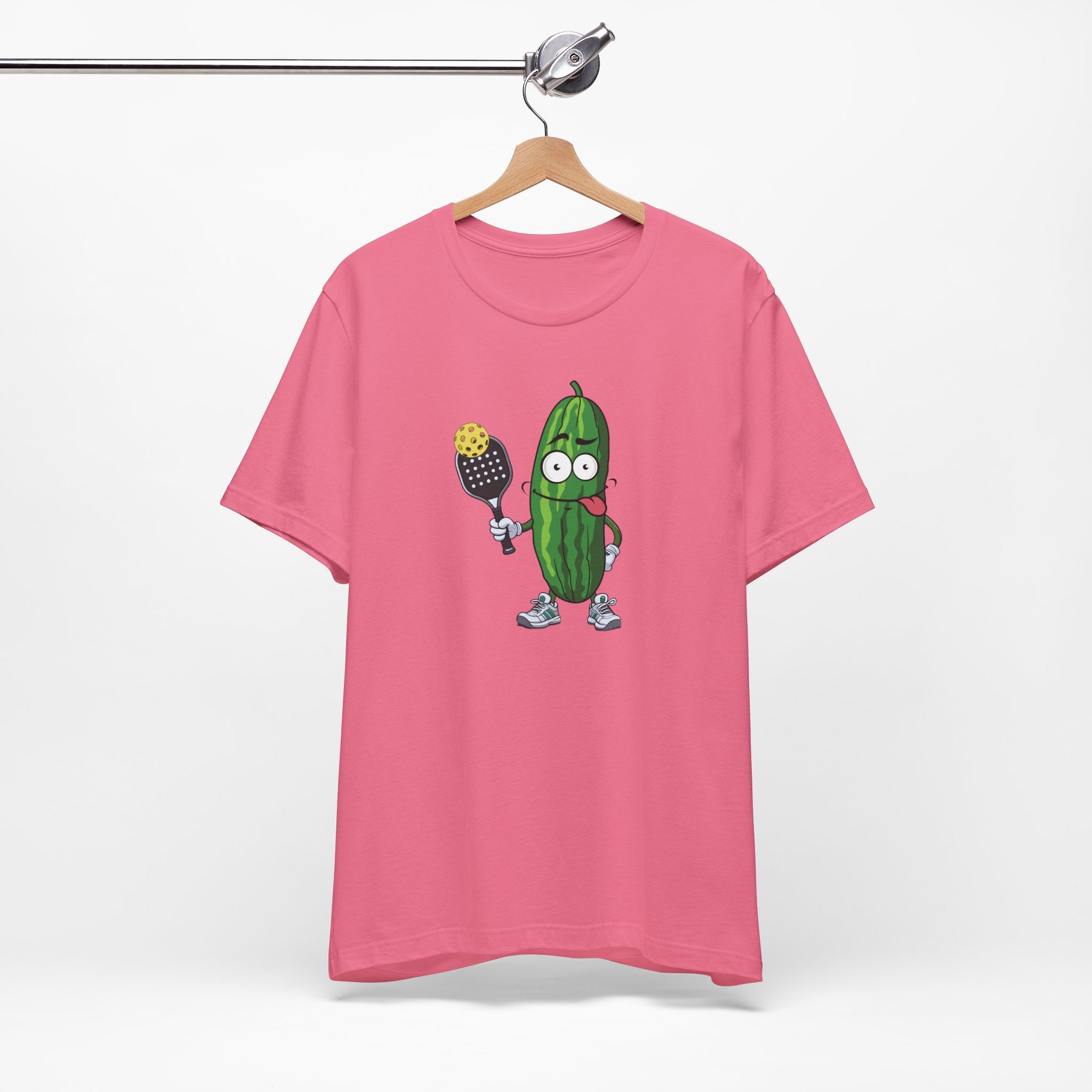 Pink t-shirt with a cartoon pickle with googly eyes and his tongue sticking out, holding a pickleball paddle with a yellow pickleball, on a hanger against a white background