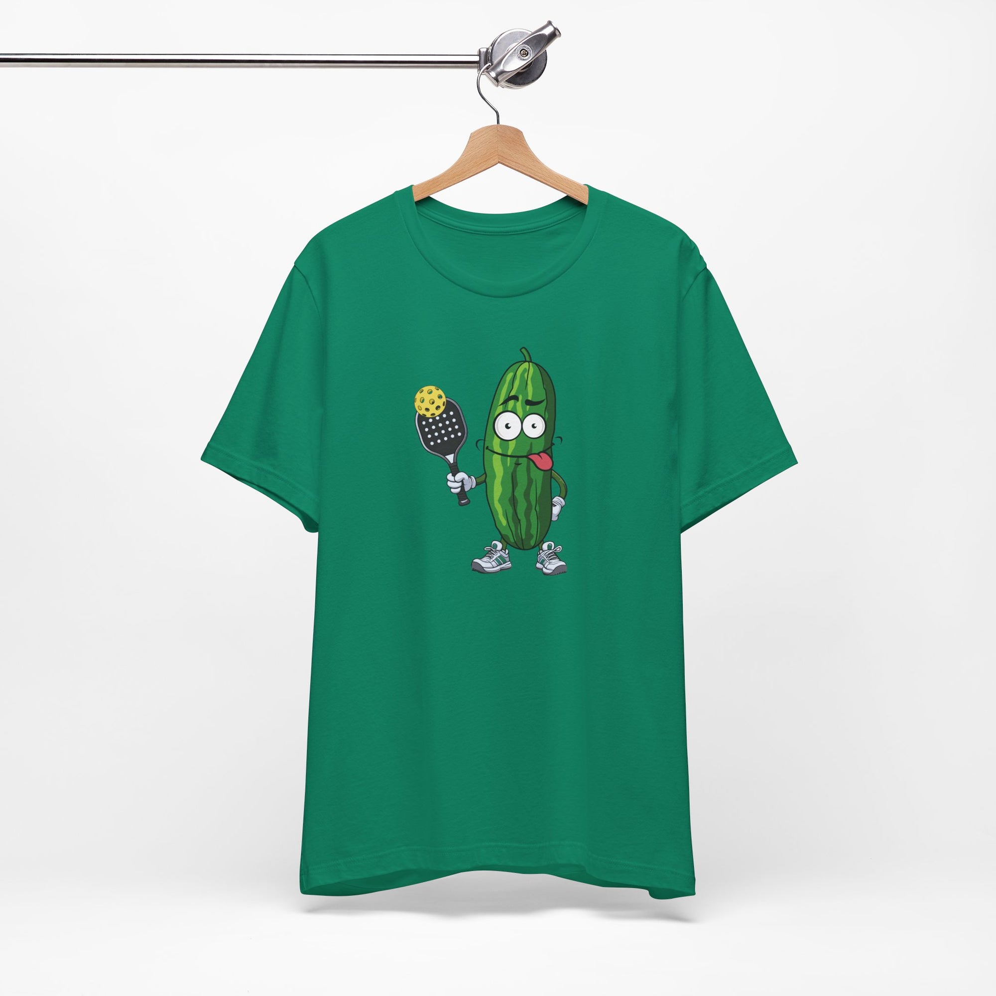 Green t-shirt with a cartoon pickle with googly eyes and his tongue sticking out, holding a pickleball paddle with a yellow pickleball, on a hanger against a white background