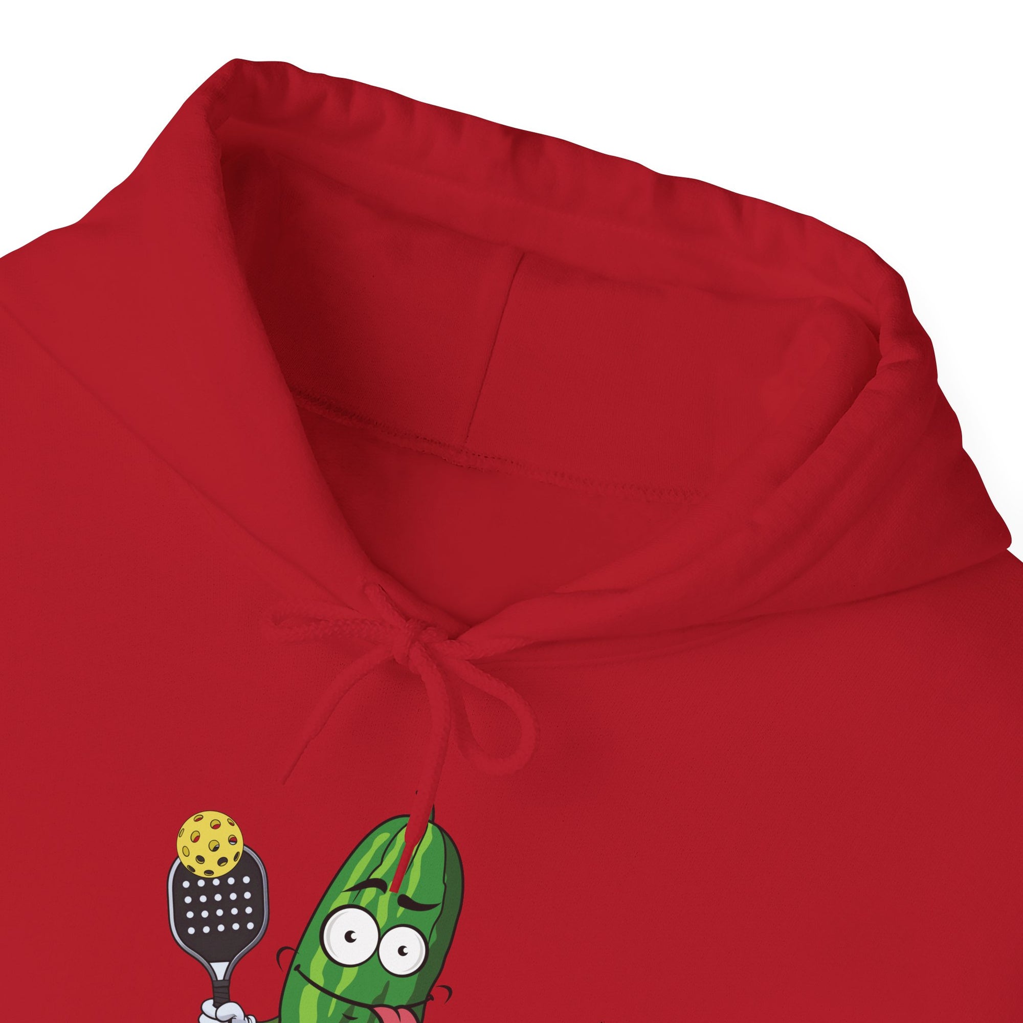 Neckline close-up of a red hoodie with a cartoon pickle with googly eyes and his tongue sticking out, holding a pickleball paddle with a yellow pickleball, against a white background