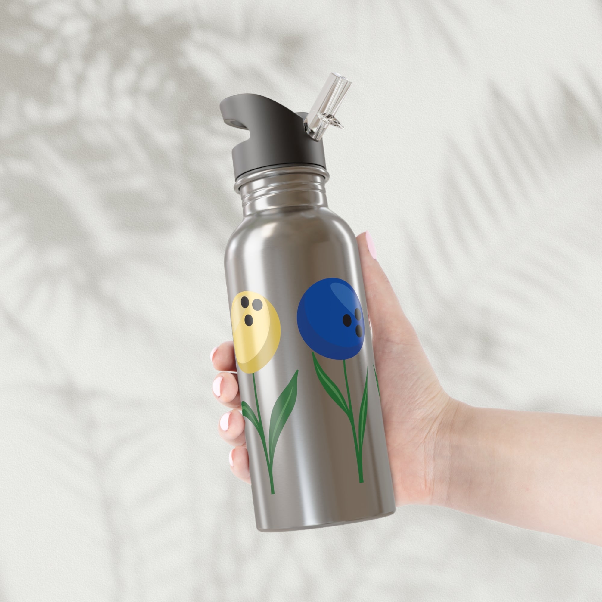 A hand holding a stainless steel water bottle with a black lid, with colorful flowers made out of bowling balls, on a white background