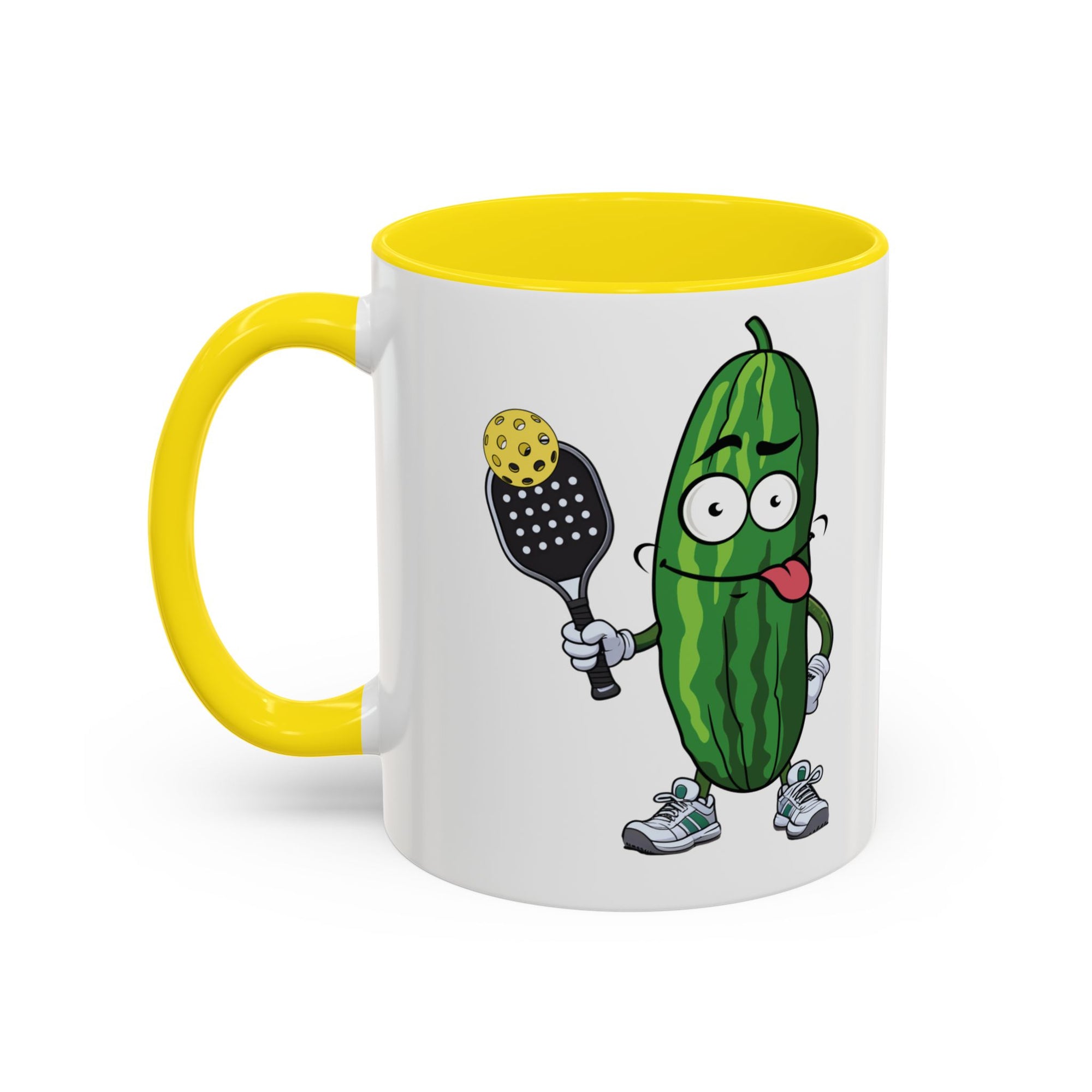 A ceramic coffee mug with yellow accents on the handle and interior, with a cartoon pickle with googly eyes and his tongue sticking out, holding a pickleball paddle with a yellow pickleball, against a white background