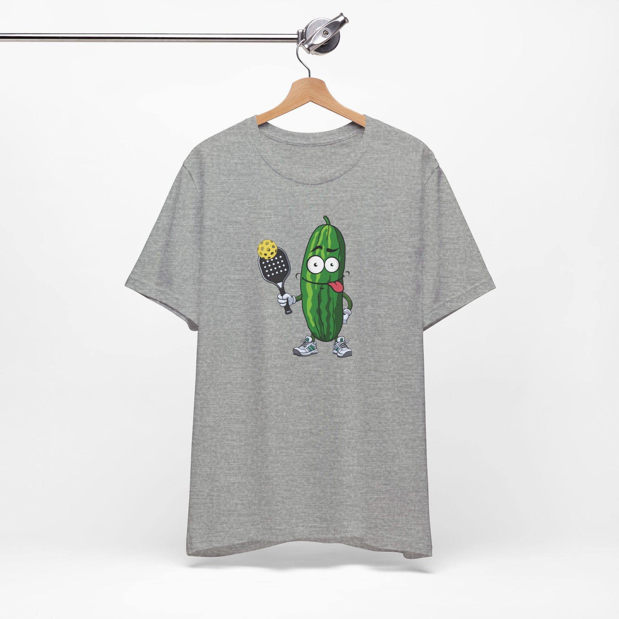 Light gray t-shirt with a cartoon pickle with googly eyes and his tongue sticking out, holding a pickleball paddle with a yellow pickleball, on a hanger against a white background