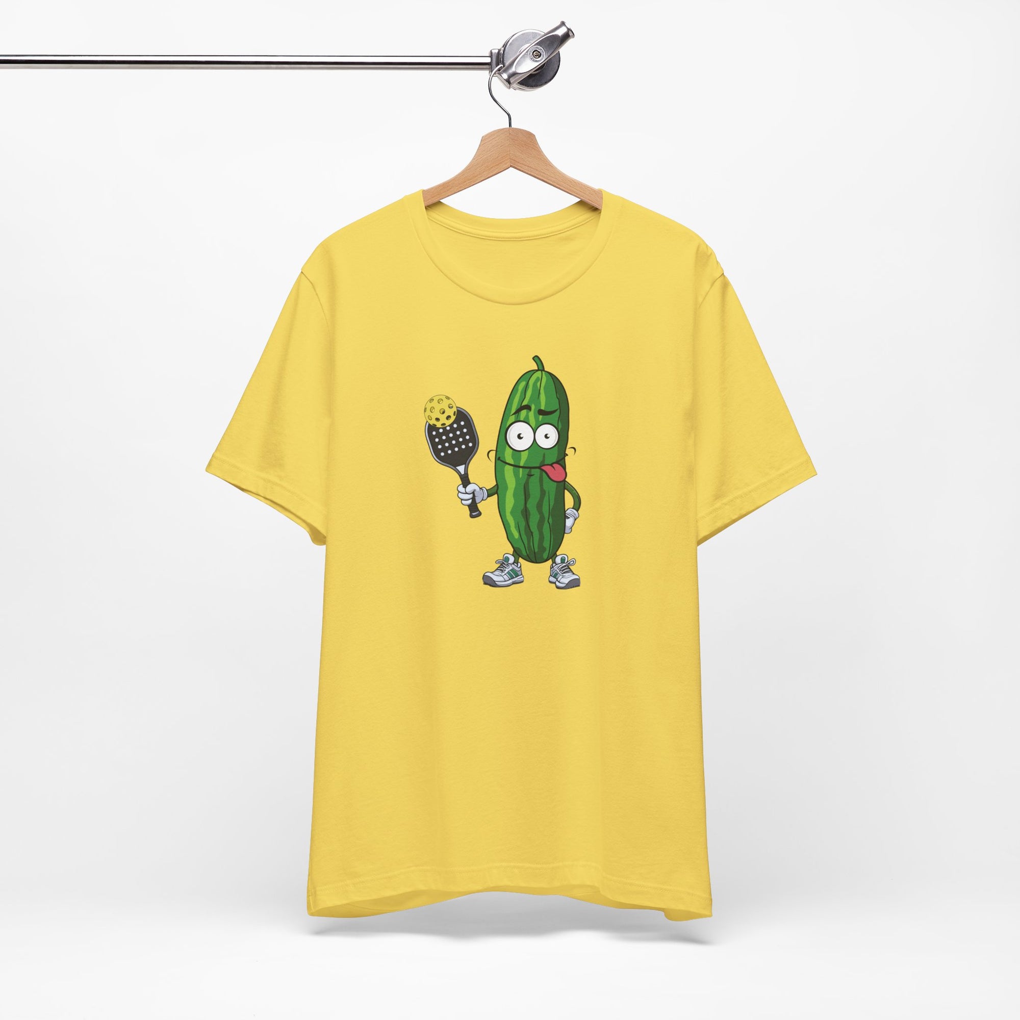 Yellow t-shirt with a cartoon pickle with googly eyes and his tongue sticking out, holding a pickleball paddle with a yellow pickleball, on a hanger against a white background