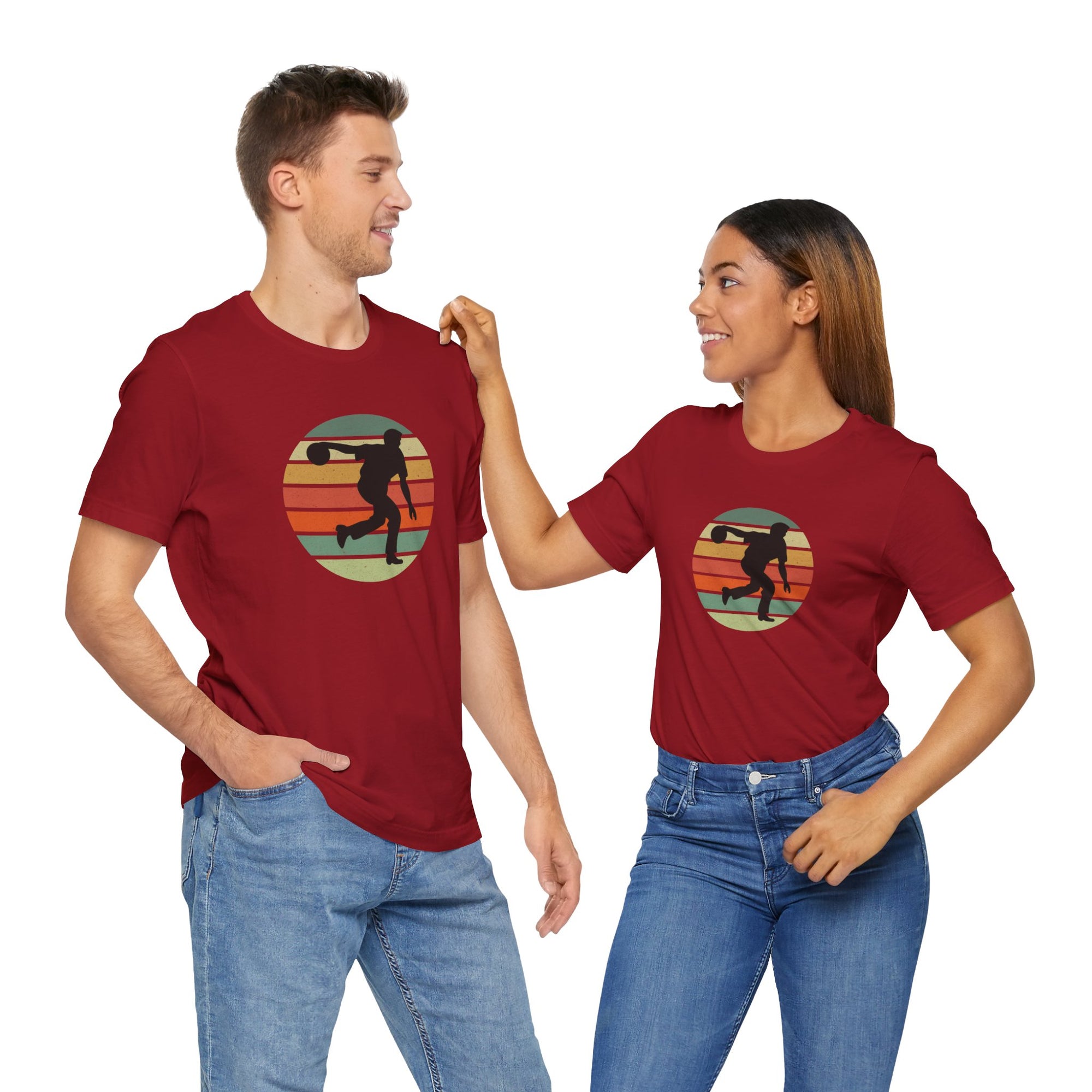 A man and a woman each wearing a red t-shirt with a graphic of a silhouette of a bowler over a colorful retro image, hanging on a wooden hanger against a white background.
