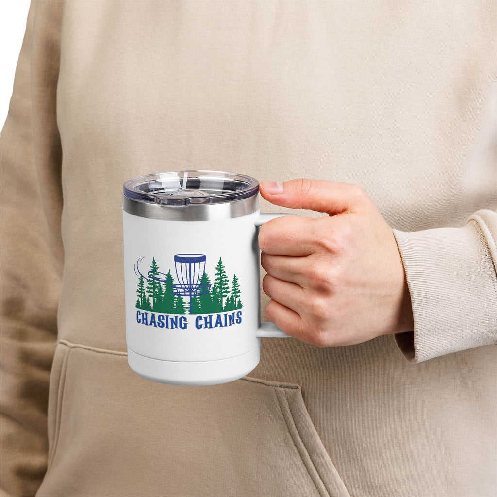A person holding a white travel coffee tumbler mug with handle and lid with 'Chasing Chains' graphic featuring a flying disc golf disc and basket and chains in front of a scenic evergreen tree landscape, against a white background