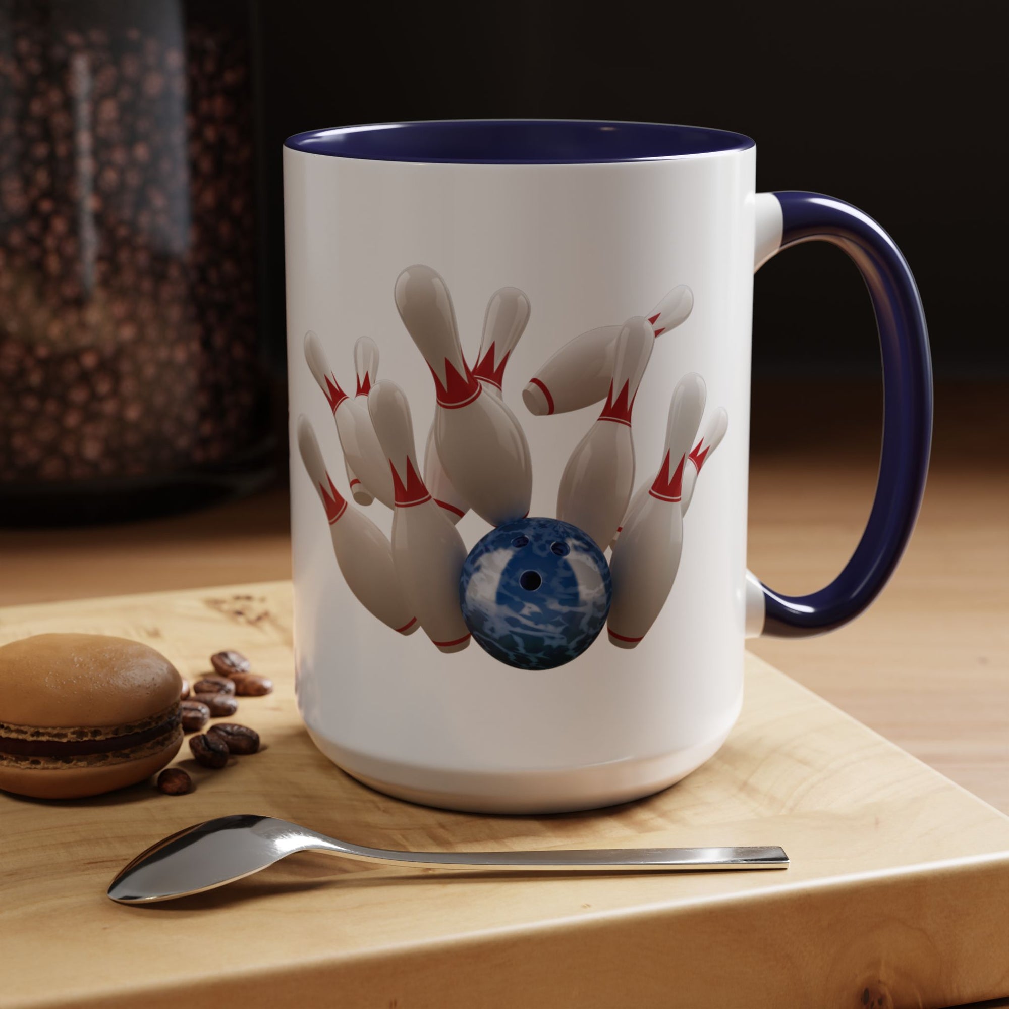 A white ceramic coffee mug with dark blue accents on the handle and interior, with a graphic of a blue bowling ball knocking over bowling pins, in a coffee shop setting