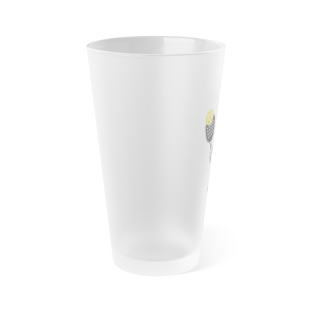 The side view of a frosted pint glass featuring an image of a cartoon pickle with googly eyes and his tongue sticking out, holding a pickleball paddle with a yellow pickleball, against a white background