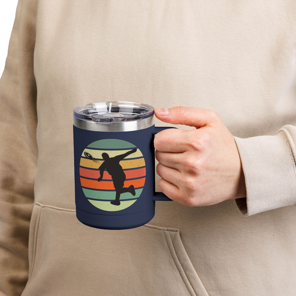Person holding a blue stainless steel coffee tumbler with handle and lid and a design on one side featuring a silhouette of a person playing disc golf over a colorful retro image, on a white background.