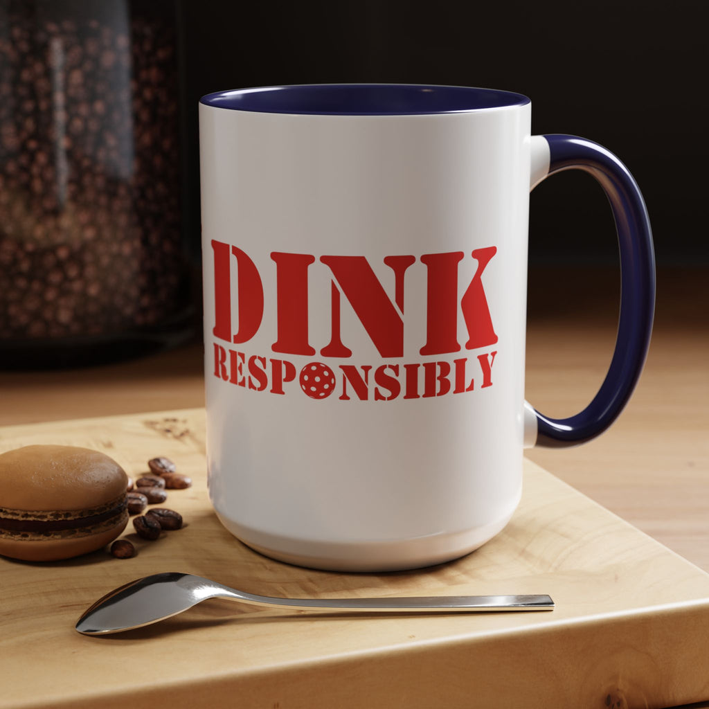 A ceramic coffee mug with dark blue accents on the handle and interior, with red text 'DINK RESPONSIBLY' with a pickleball replacing the "O" in "Responsibly" in a coffee shop setting