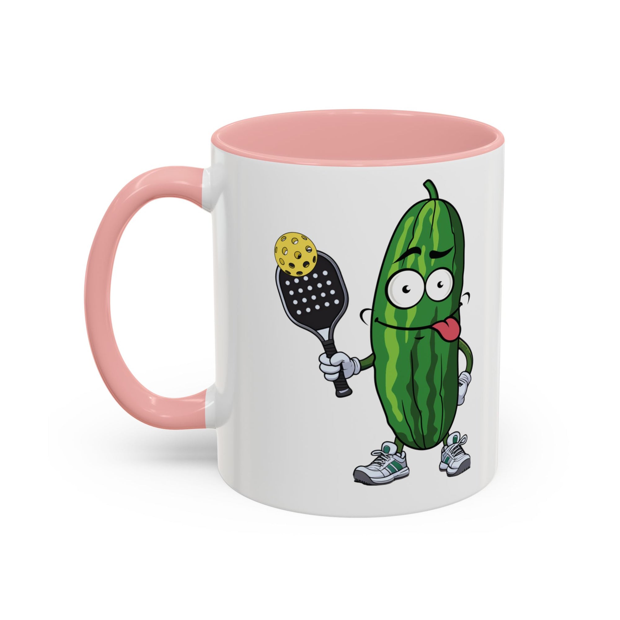 A ceramic coffee mug with pink accents on the handle and interior, with a cartoon pickle with googly eyes and his tongue sticking out, holding a pickleball paddle with a yellow pickleball, against a white background