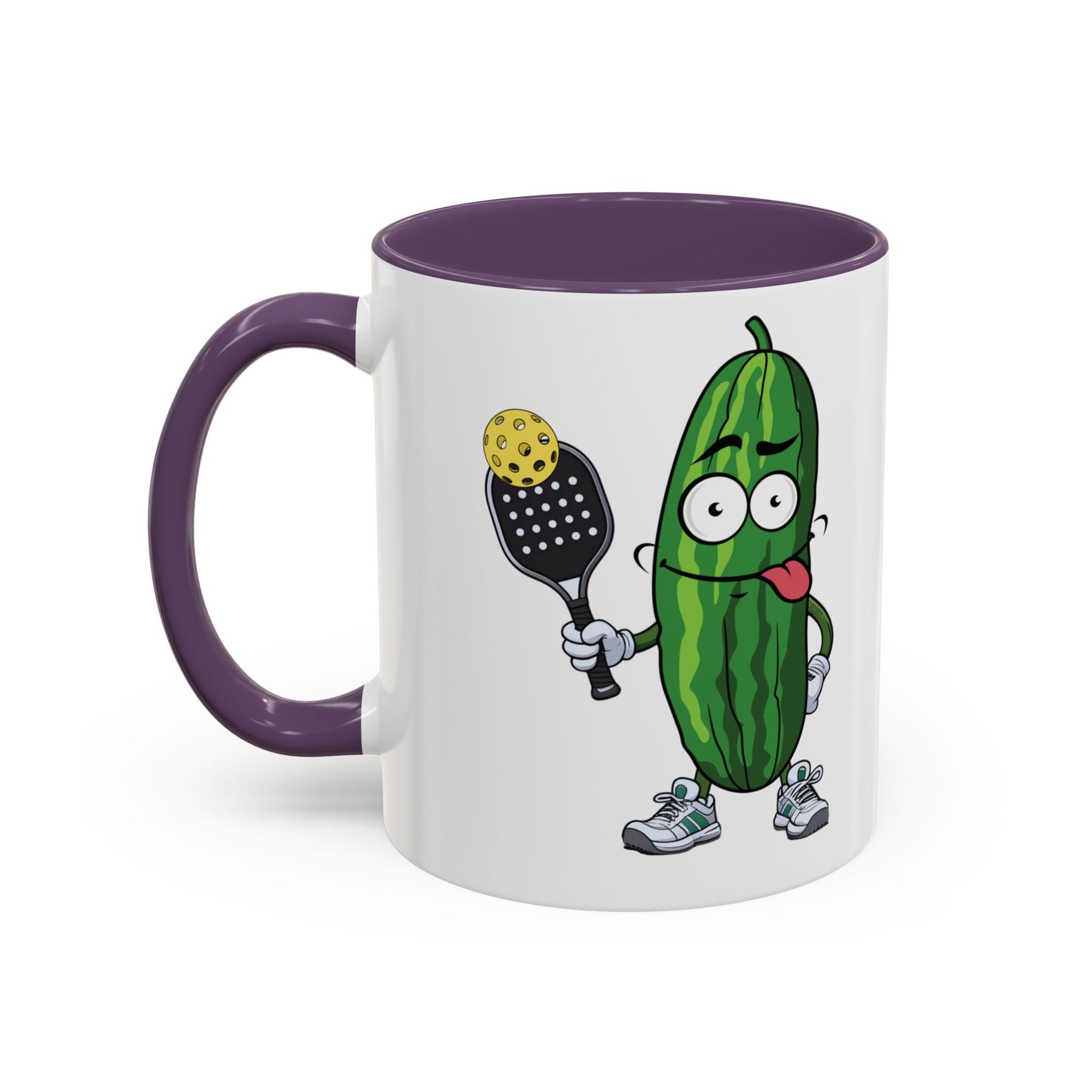 A ceramic coffee mug with purple accents on the handle and interior, with a cartoon pickle with googly eyes and his tongue sticking out, holding a pickleball paddle with a yellow pickleball, against a white background