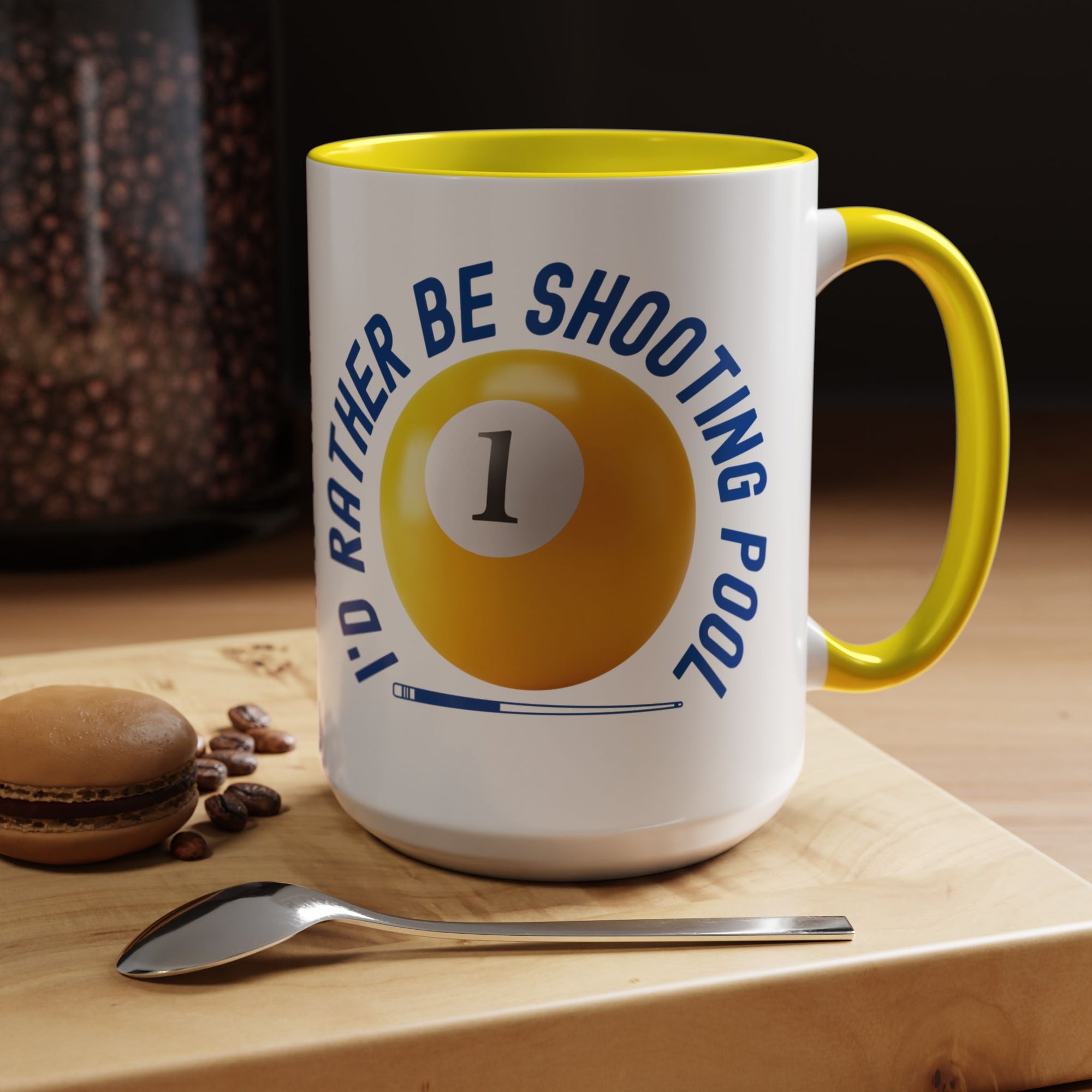 Ceramic coffee mug with yellow accents on the handle and interior, with a yellow billiards ball and cue stick graphic and the text "I'd Rather Be Shooting Pool" in a coffee house setting.