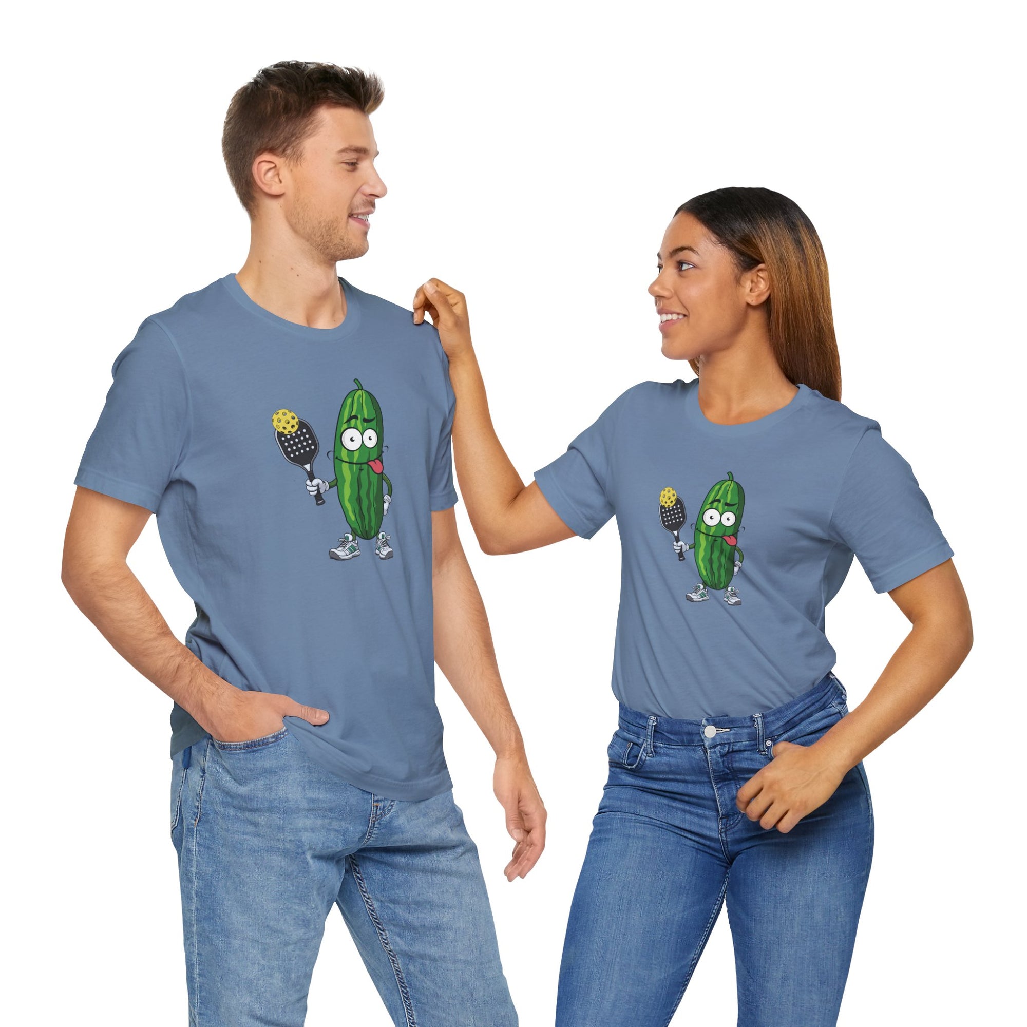 A man and a woman both wearing a steel blue t-shirt with a cartoon pickle with googly eyes and his tongue sticking out, holding a pickleball paddle with a yellow pickleball, against a white background