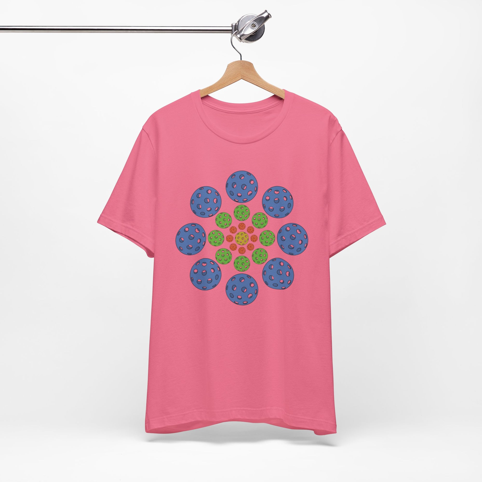 Pink t-shirt with colorful geometric design of pickleballs on a hanger against a white background