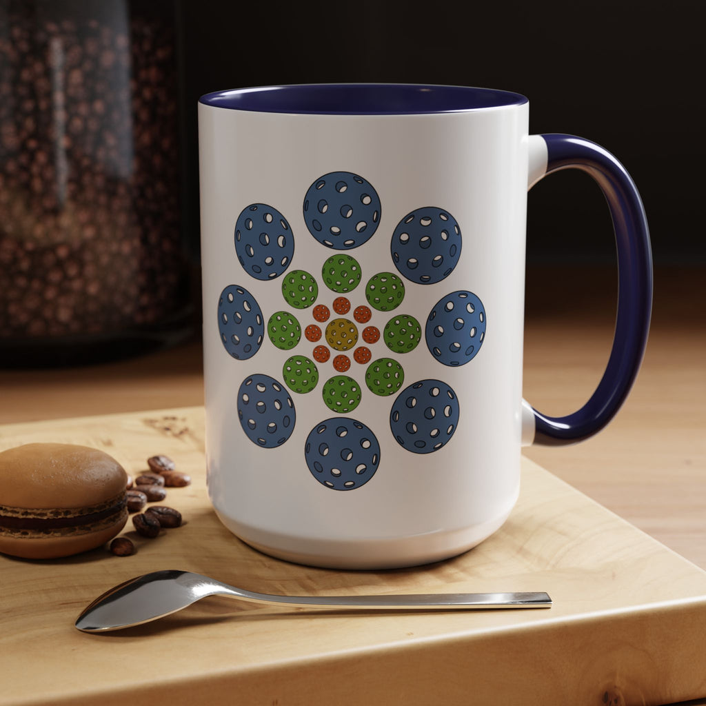 A ceramic coffee mug with dark blue accents on the handle and interior, with a colorful geometric design of pickleballs set in a coffee shop setting