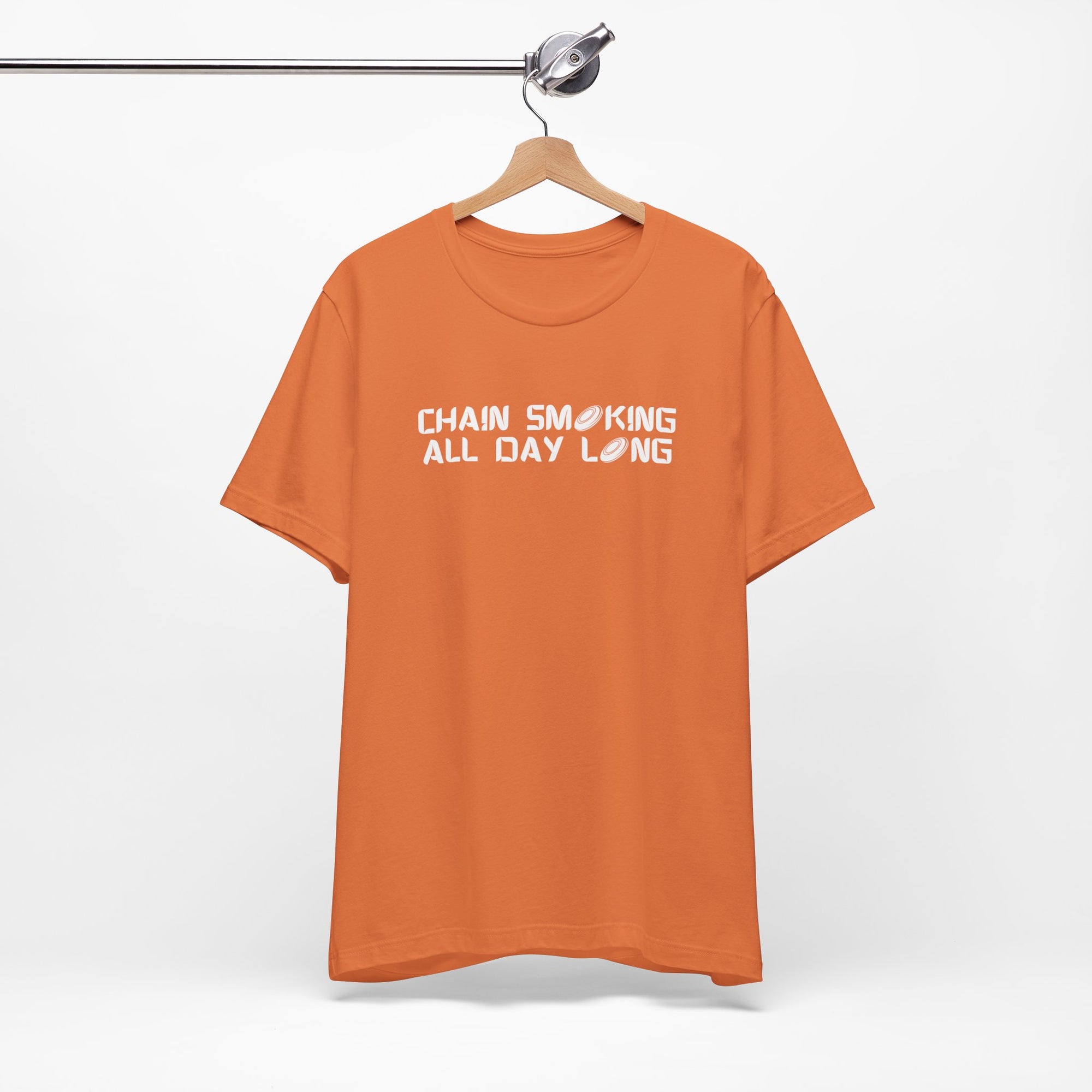 Orange t-shirt with 'CHAIN SMOKING ALL DAY LONG' text with the O's replaced by disc golf discs, on a hanger against a white background