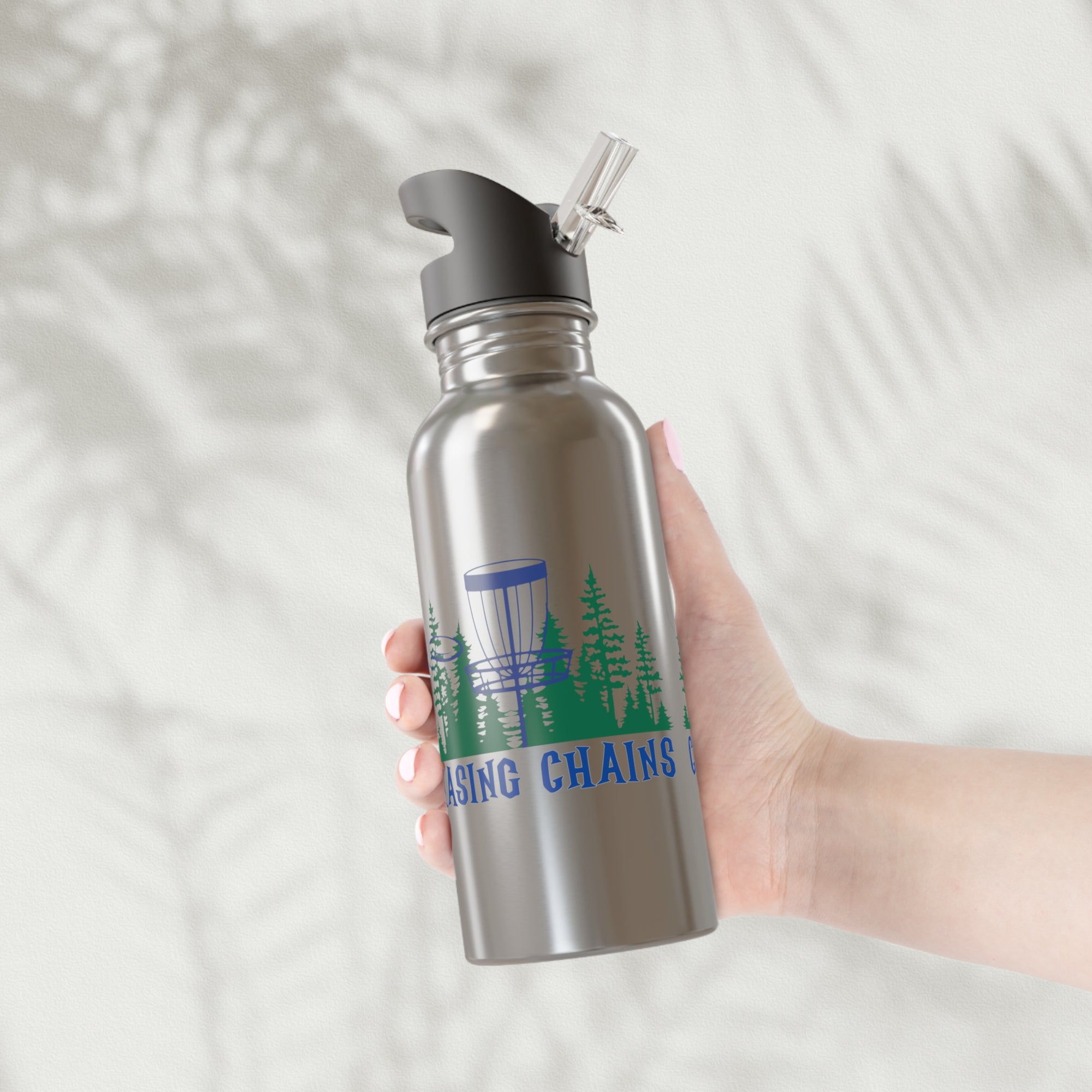 A hand holding a stainless steel water bottle with lid, with 'Chasing Chains' graphic featuring a flying disc golf disc and basket and chains in front of a scenic evergreen tree landscape, against a white background