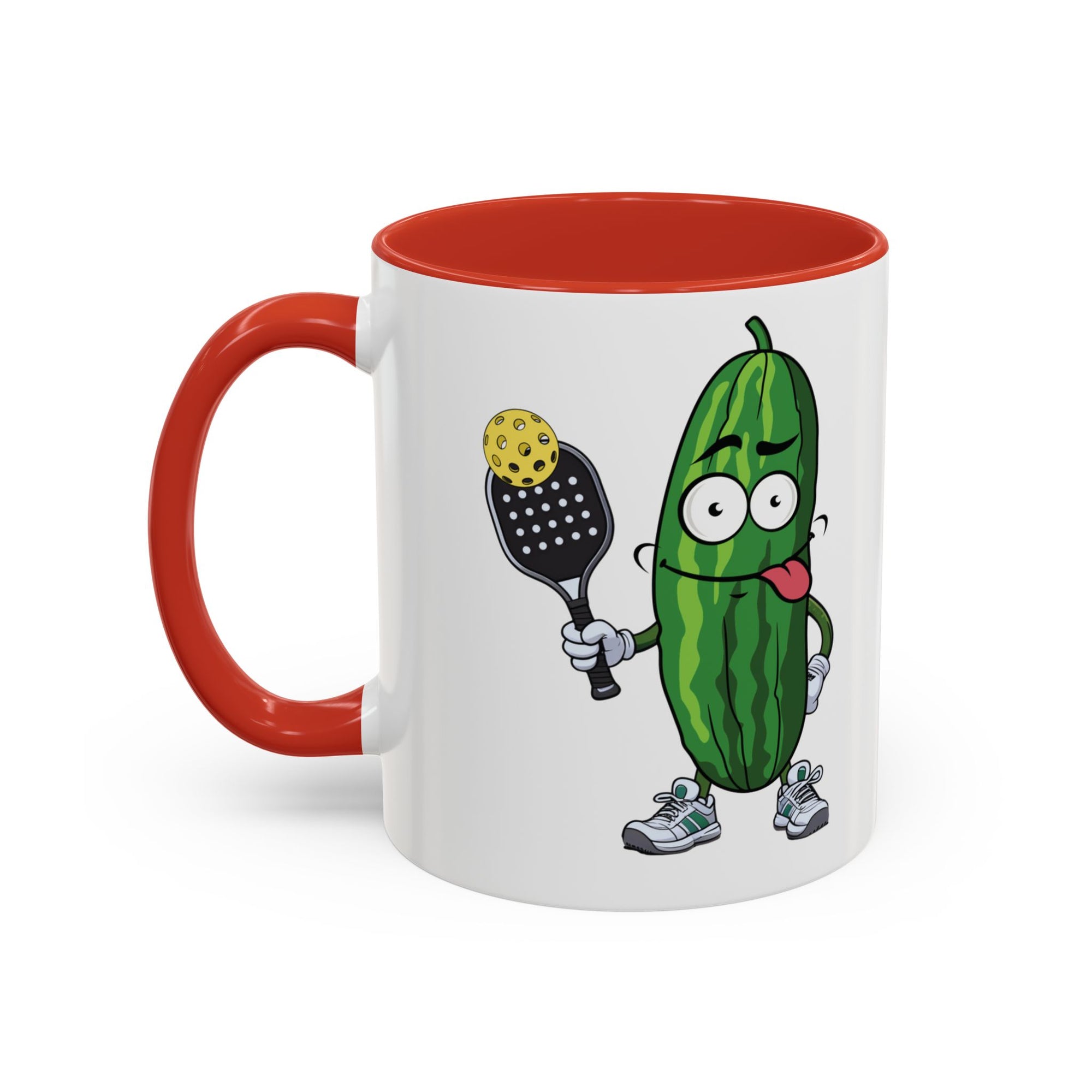 A ceramic coffee mug with red accents on the handle and interior, with a cartoon pickle with googly eyes and his tongue sticking out, holding a pickleball paddle with a yellow pickleball, against a white background